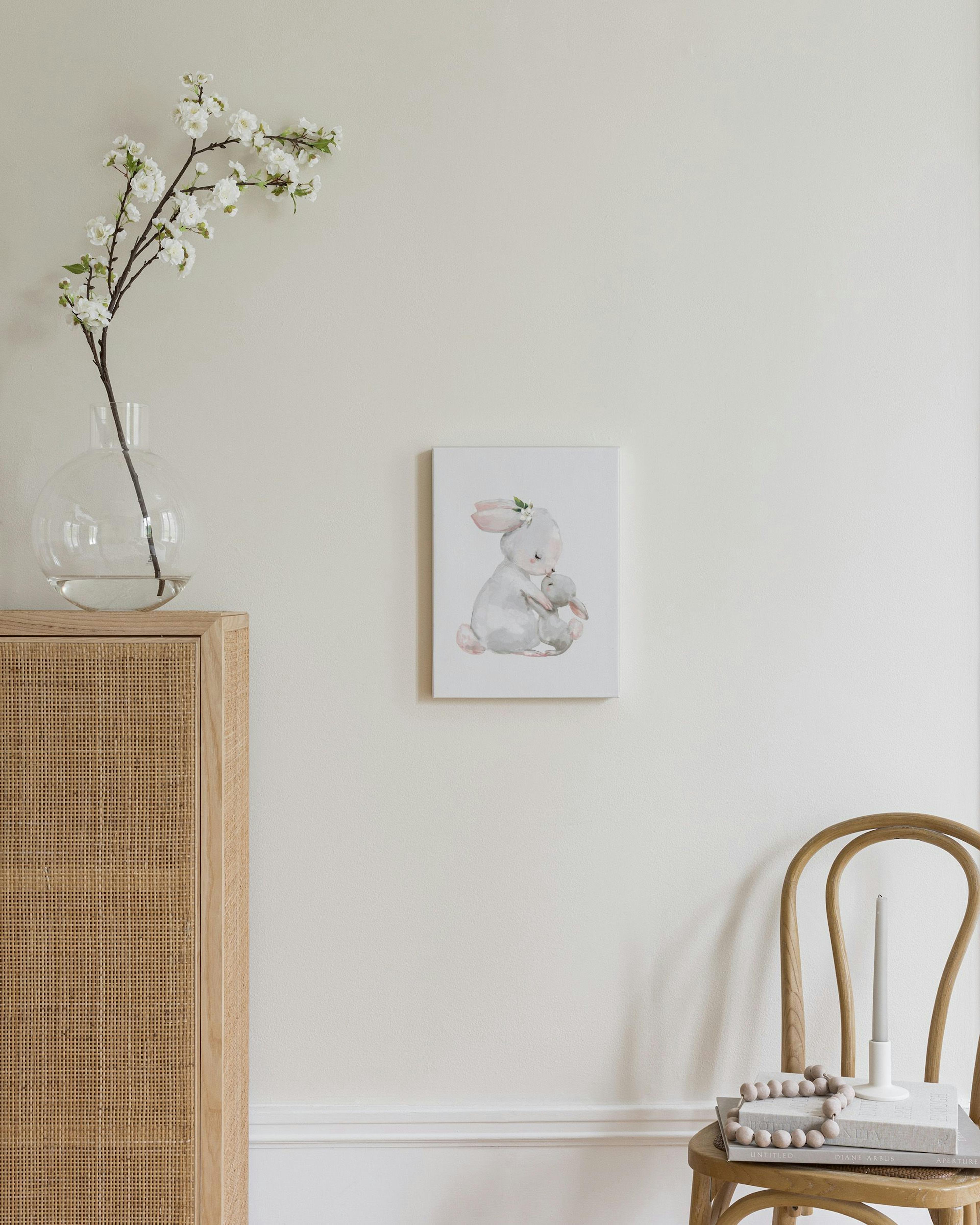 A canvas of a mother rabbit hugging a baby bunny, displayed above a wooden dresser with a lamp and vase.