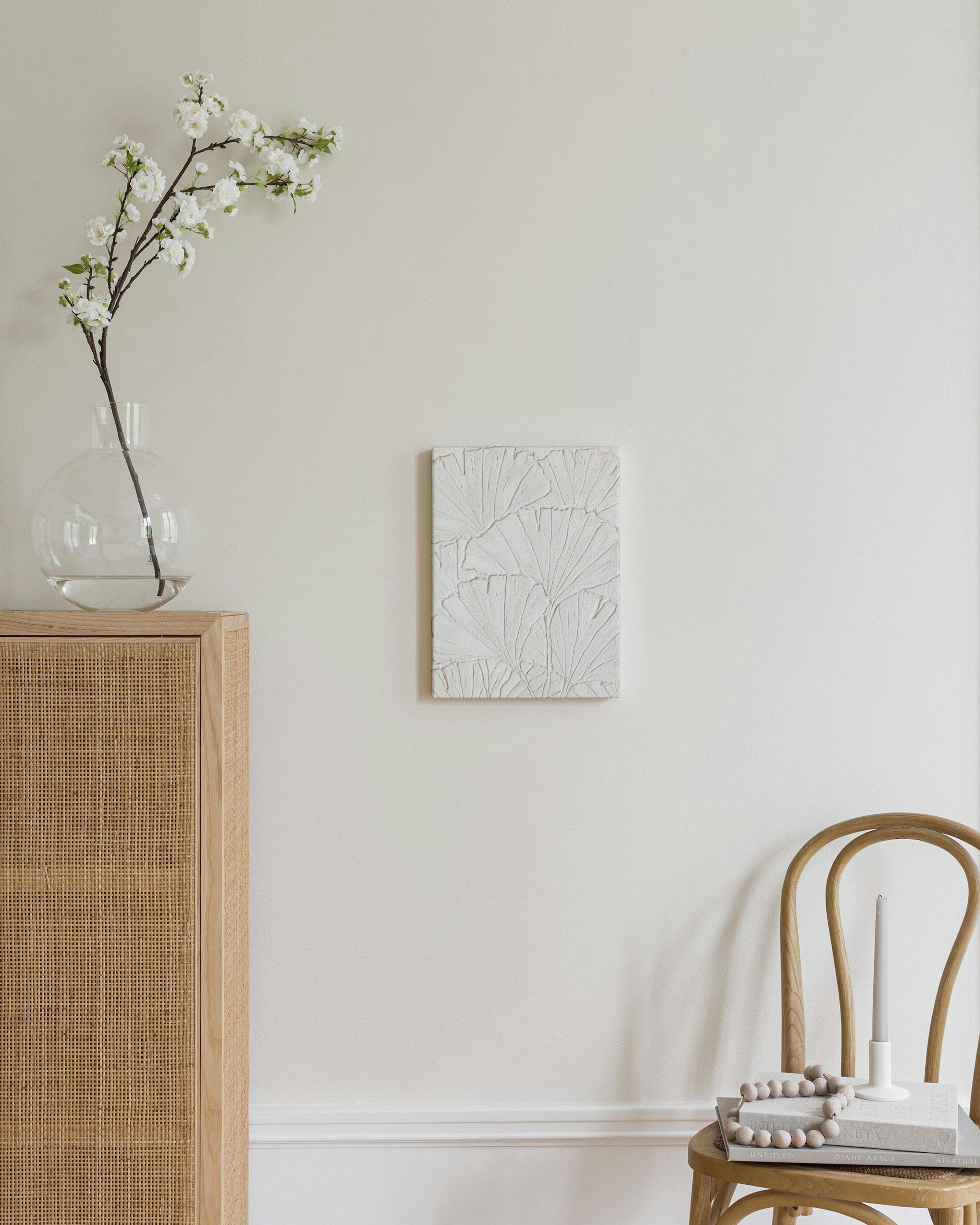 Lienzo blanco texturizado con diseño de hojas de ginkgo, colgado en pared clara sobre mueble de madera y silla.