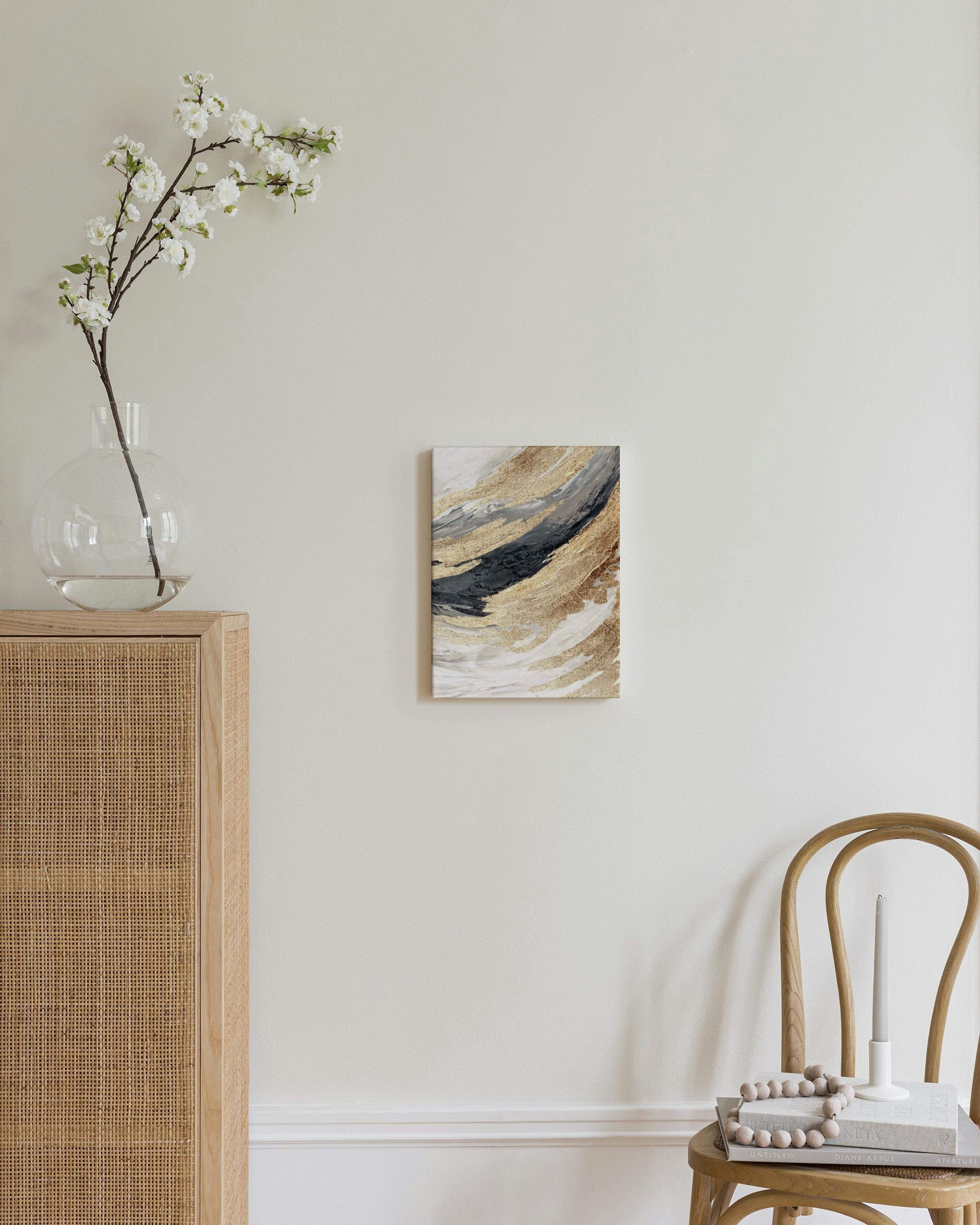 A canvas with abstract gold, gray, and black brushstrokes, hanging on a light wall in a room with a wooden cabinet and chair.