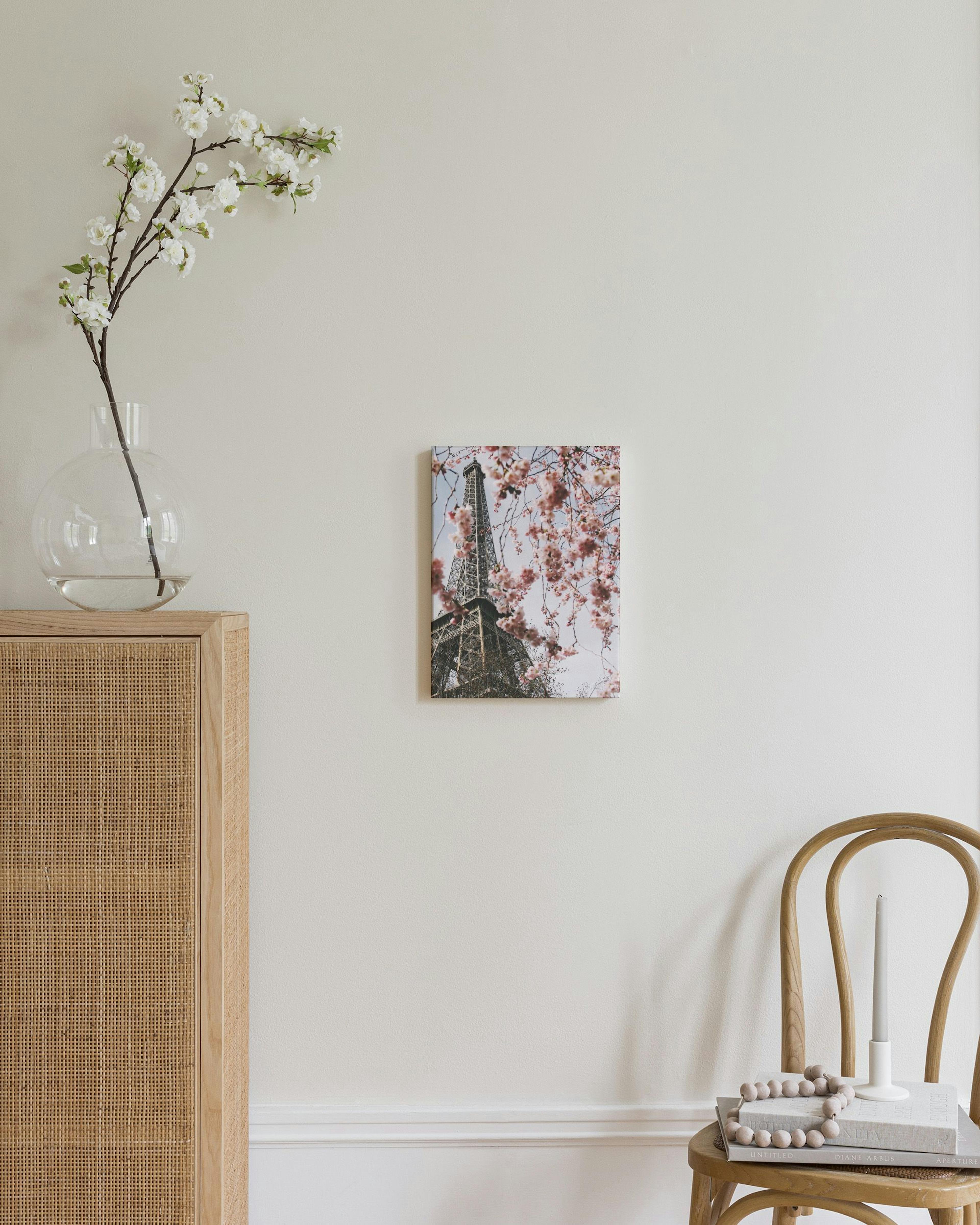 A canvas displaying the Eiffel Tower framed by pink cherry blossoms, hanging on a beige wall in a calm room.