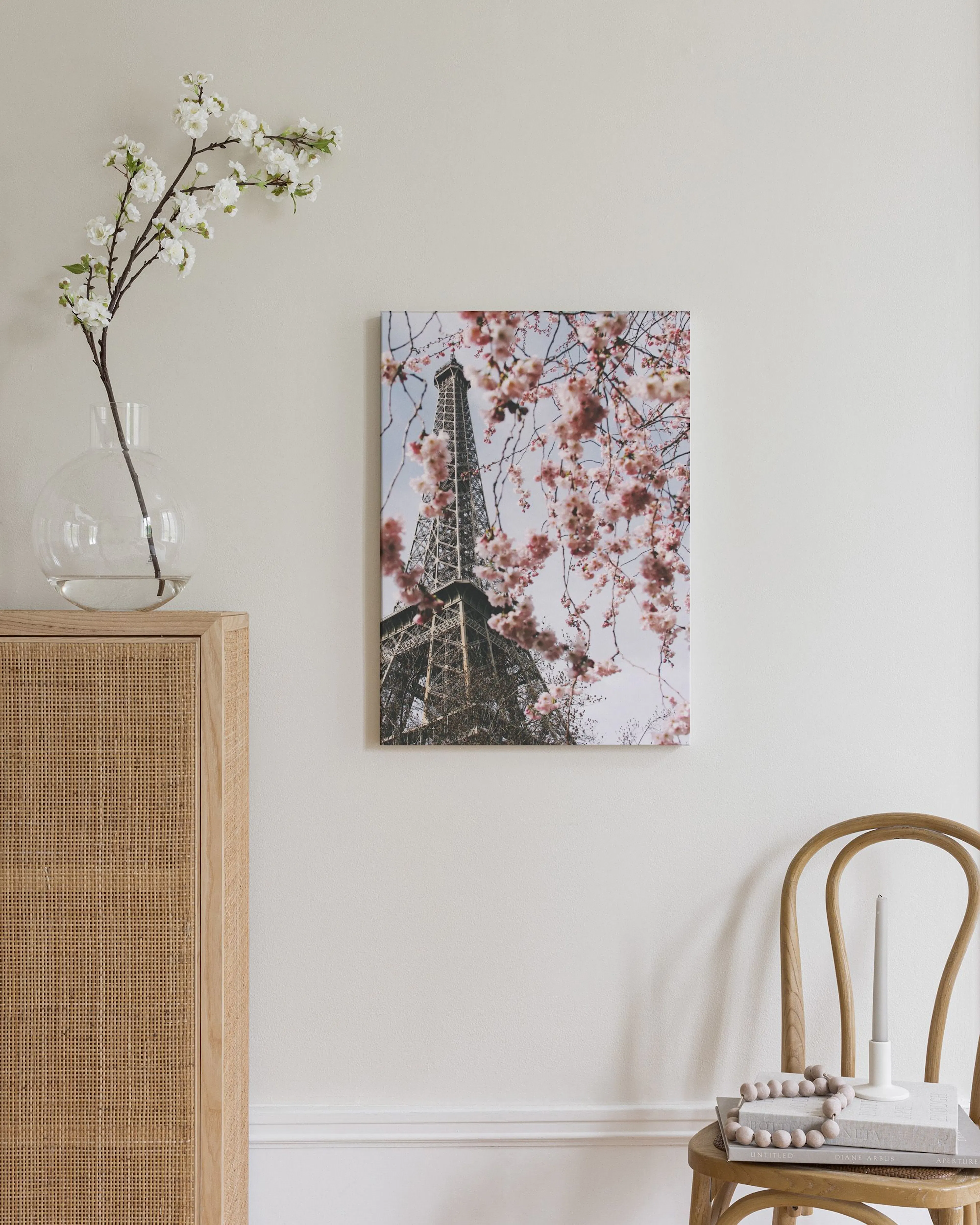 Lærred med Eiffeltårnet og lyserøde kirsebærblomster ophængt i et lyst rum med en hvid vase og puf.