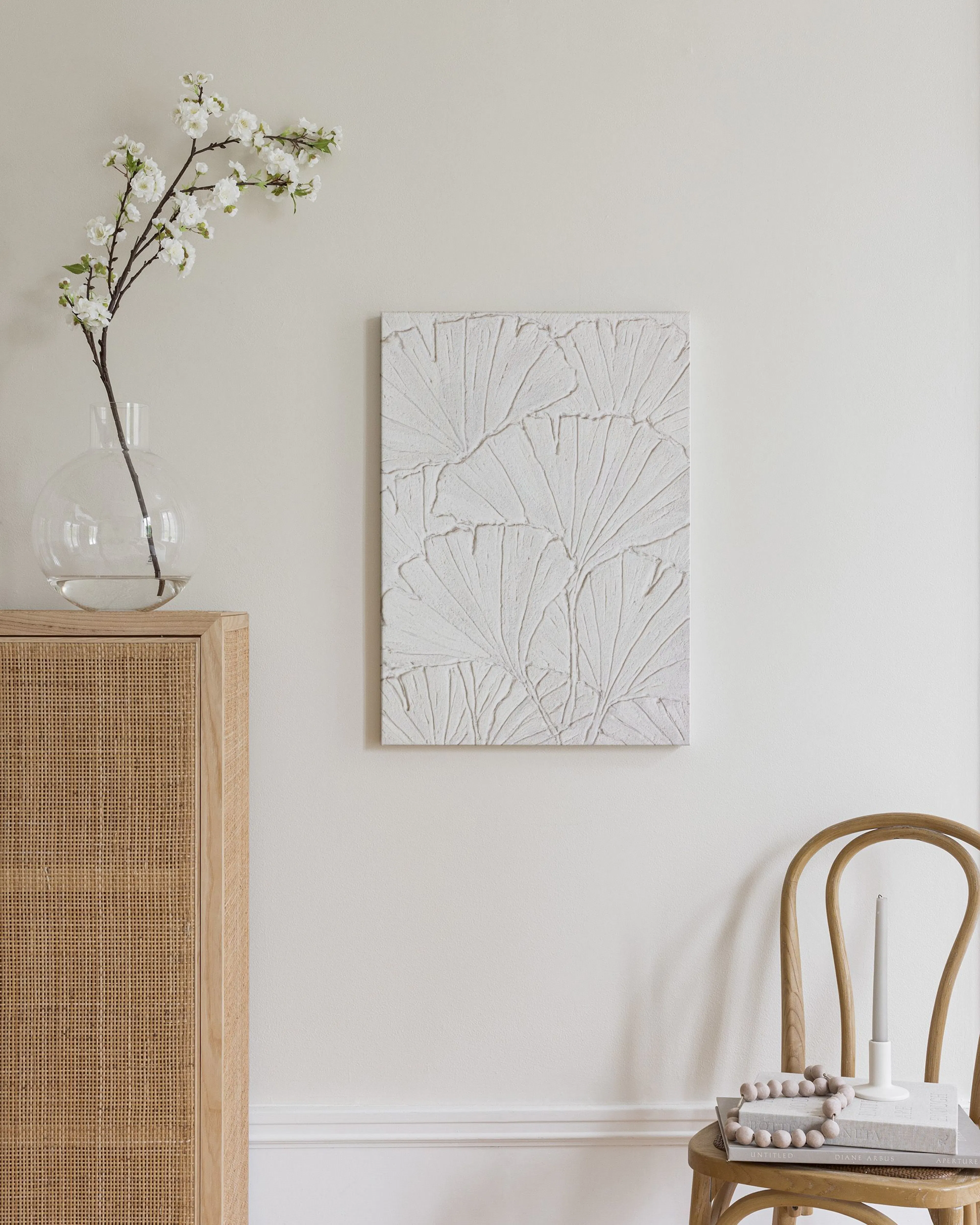 A canvas with a textured white ginkgo leaf pattern, hanging on a cream wall next to a wooden cabinet and chair.