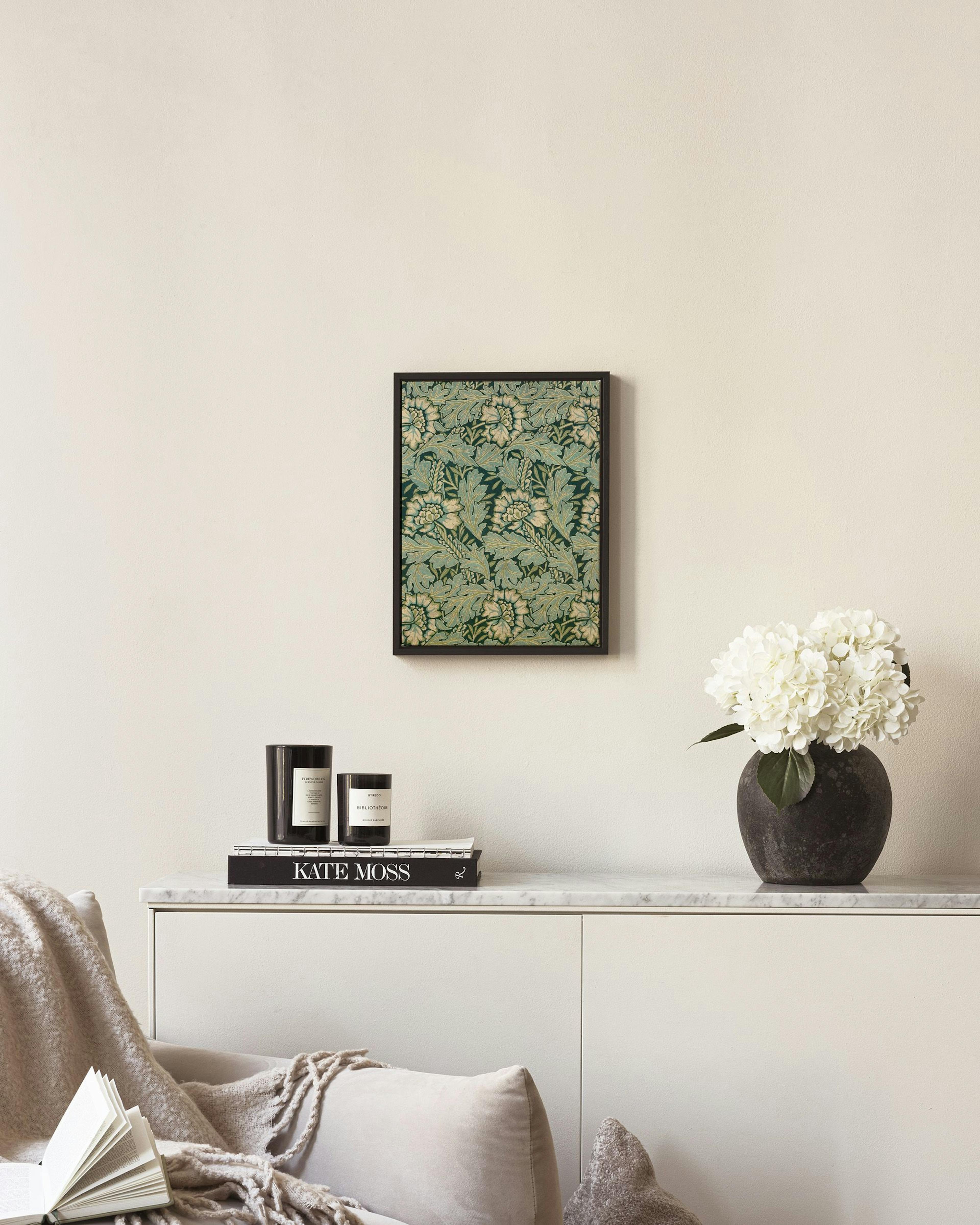A canvas with a dark green and teal floral pattern, hanging on a light wall above a wooden chair with a candle.