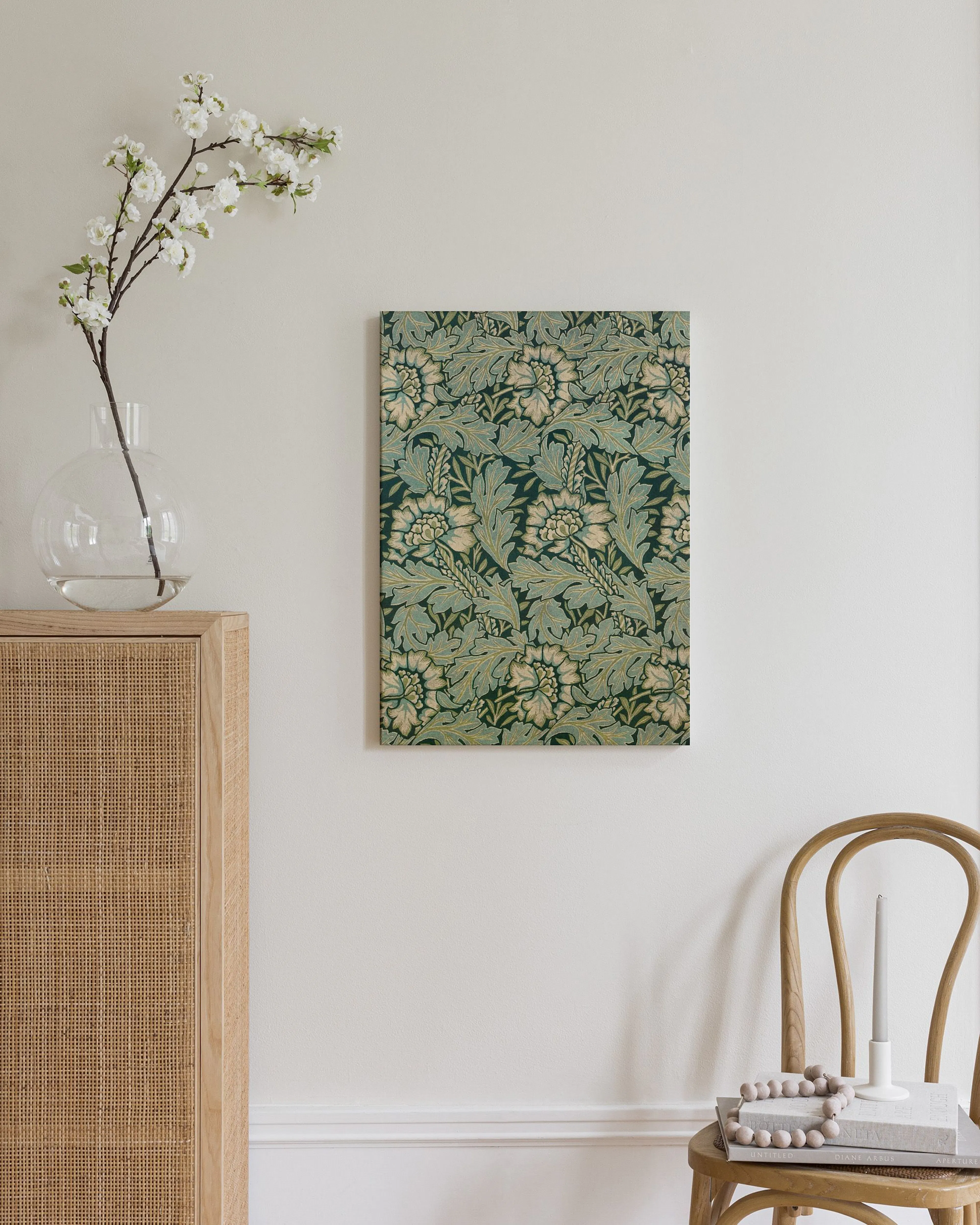 A canvas with a dark green and light teal botanical pattern, hanging above a woven cabinet with a vase of white flowers.