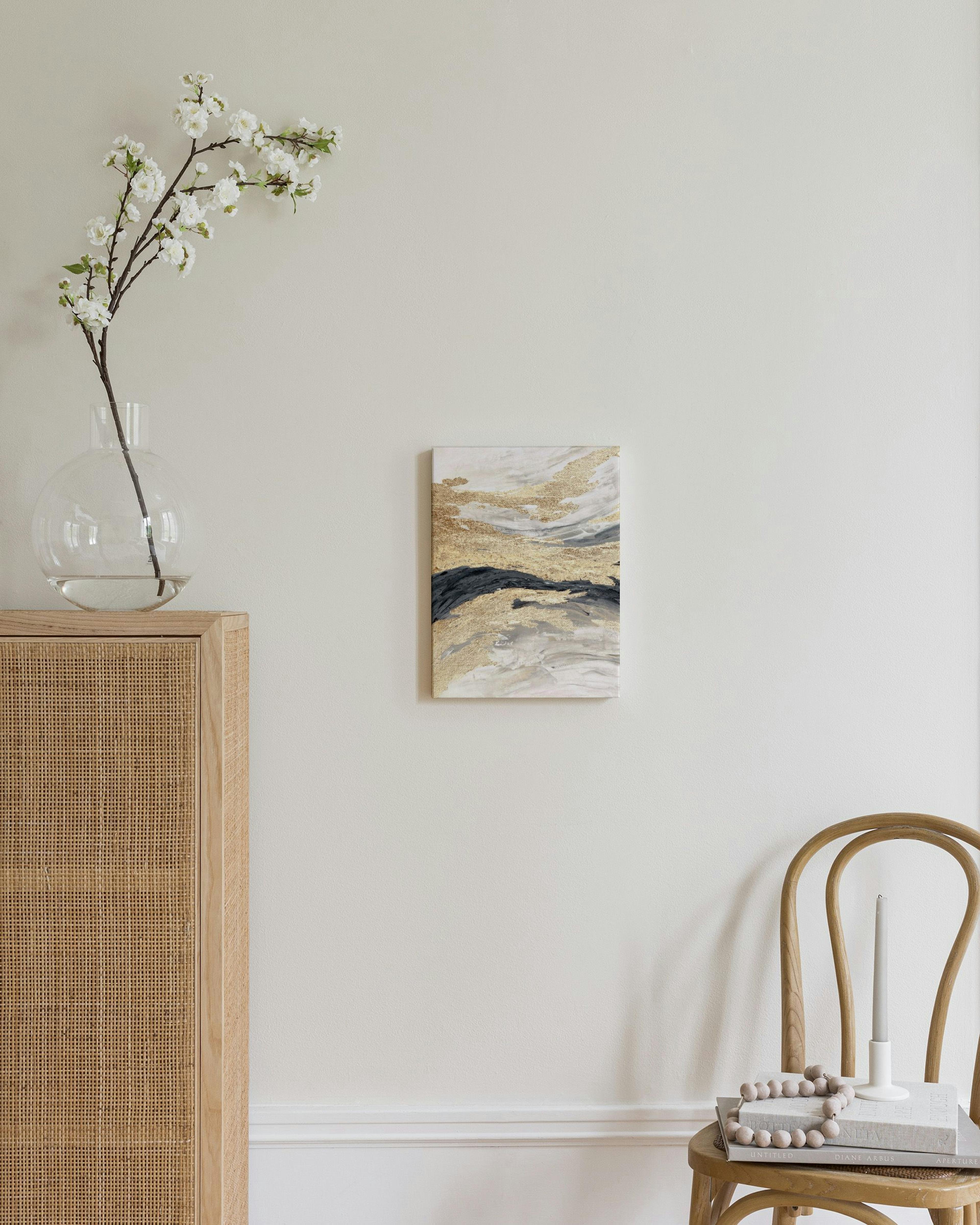 Toile abstraite aux tons dorés, gris et noirs, accrochée au-dessus dune chaise en bois dans un intérieur moderne.