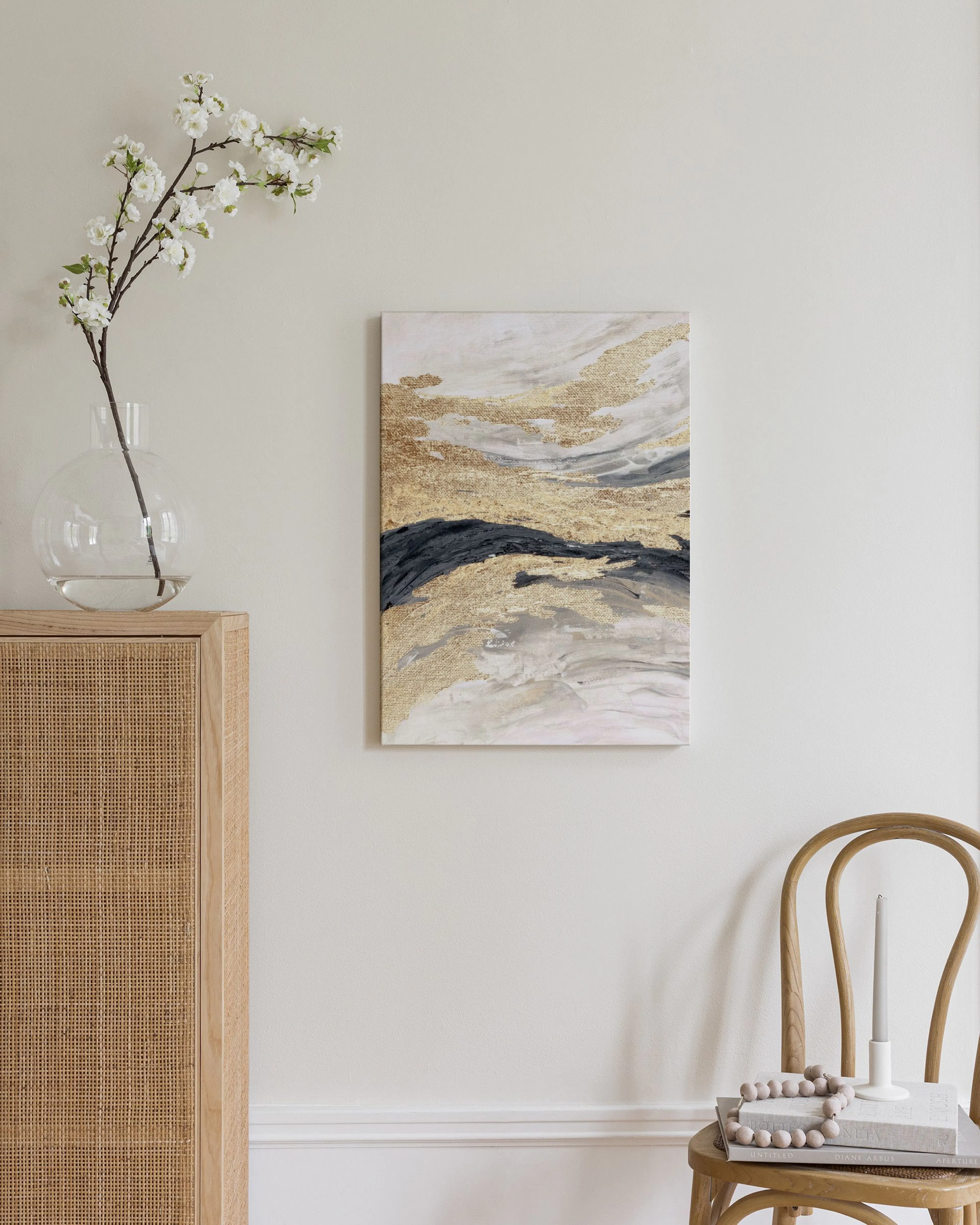 Lienzo abstracto con texturas doradas y pinceladas en gris y negro, colgado sobre un mueble de madera y una silla.