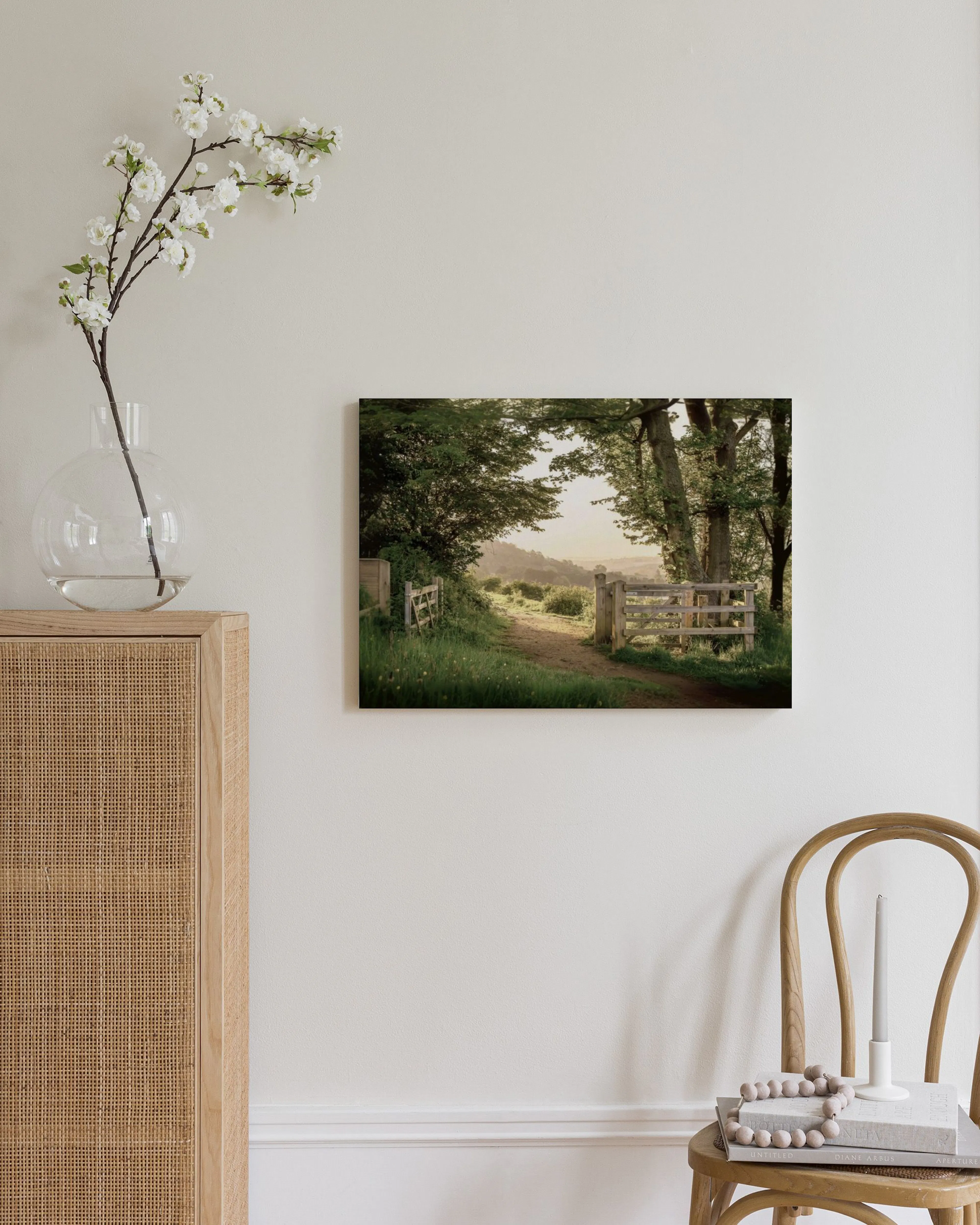 A canvas displaying a serene landscape with a dirt path, wooden fence, and trees, hanging on a light wall next to a wooden cabin