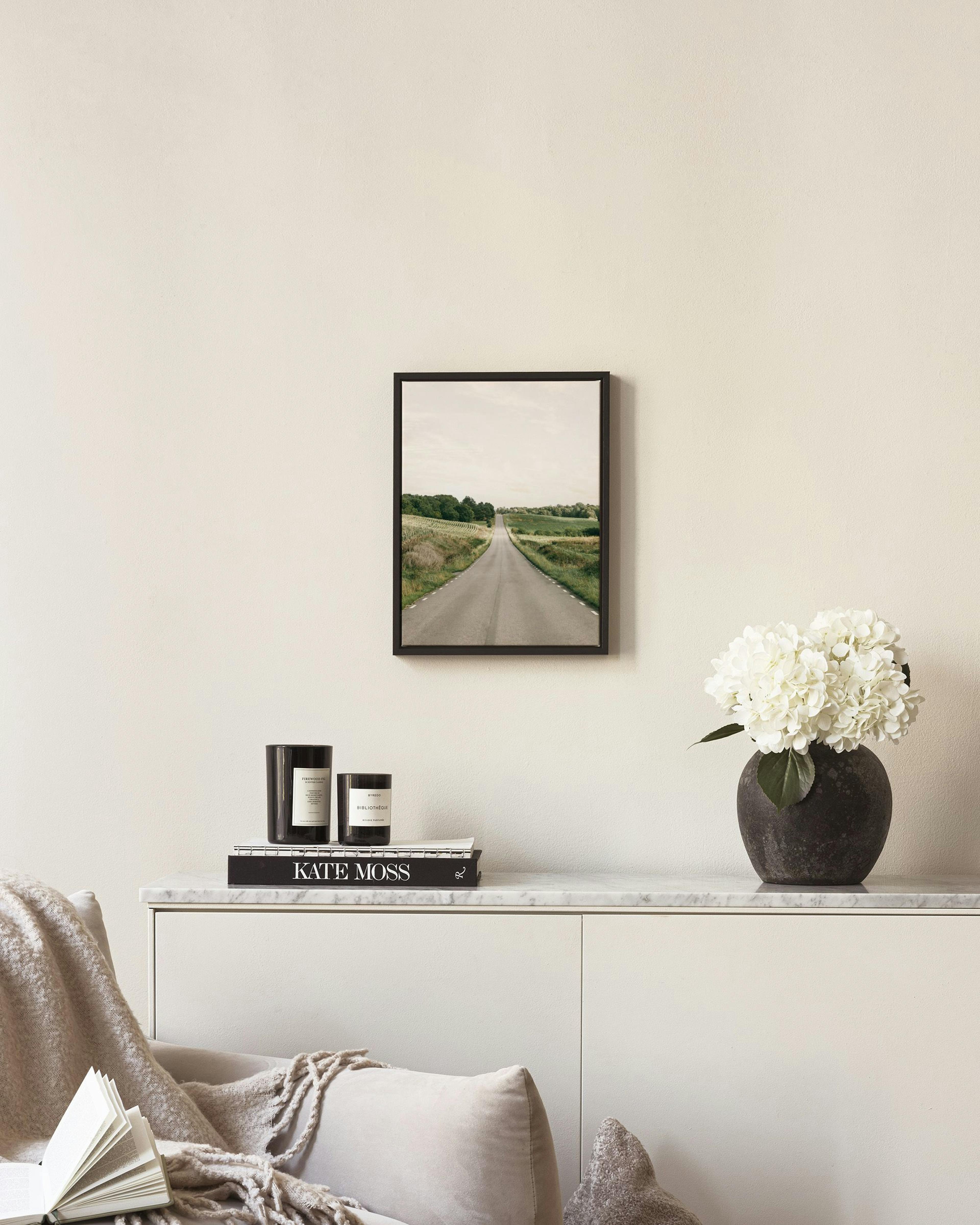 A canvas of a long, straight road extending through green fields under a light sky, hanging on a white wall above a wooden chair
