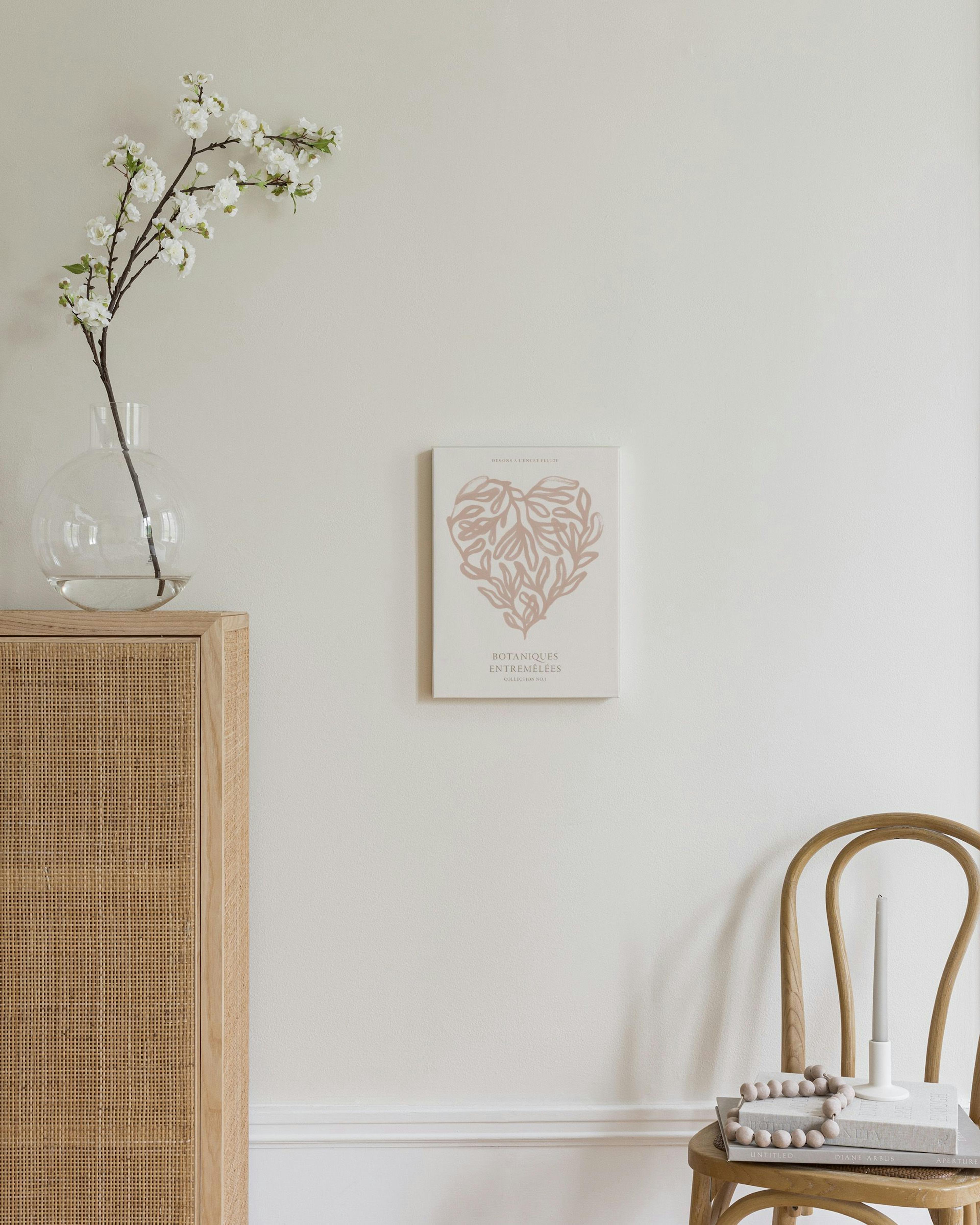Lienzo con diseño de corazón formado por hojas botánicas en tonos rosa pálido, colgado en una pared clara junto a un jarrón y un