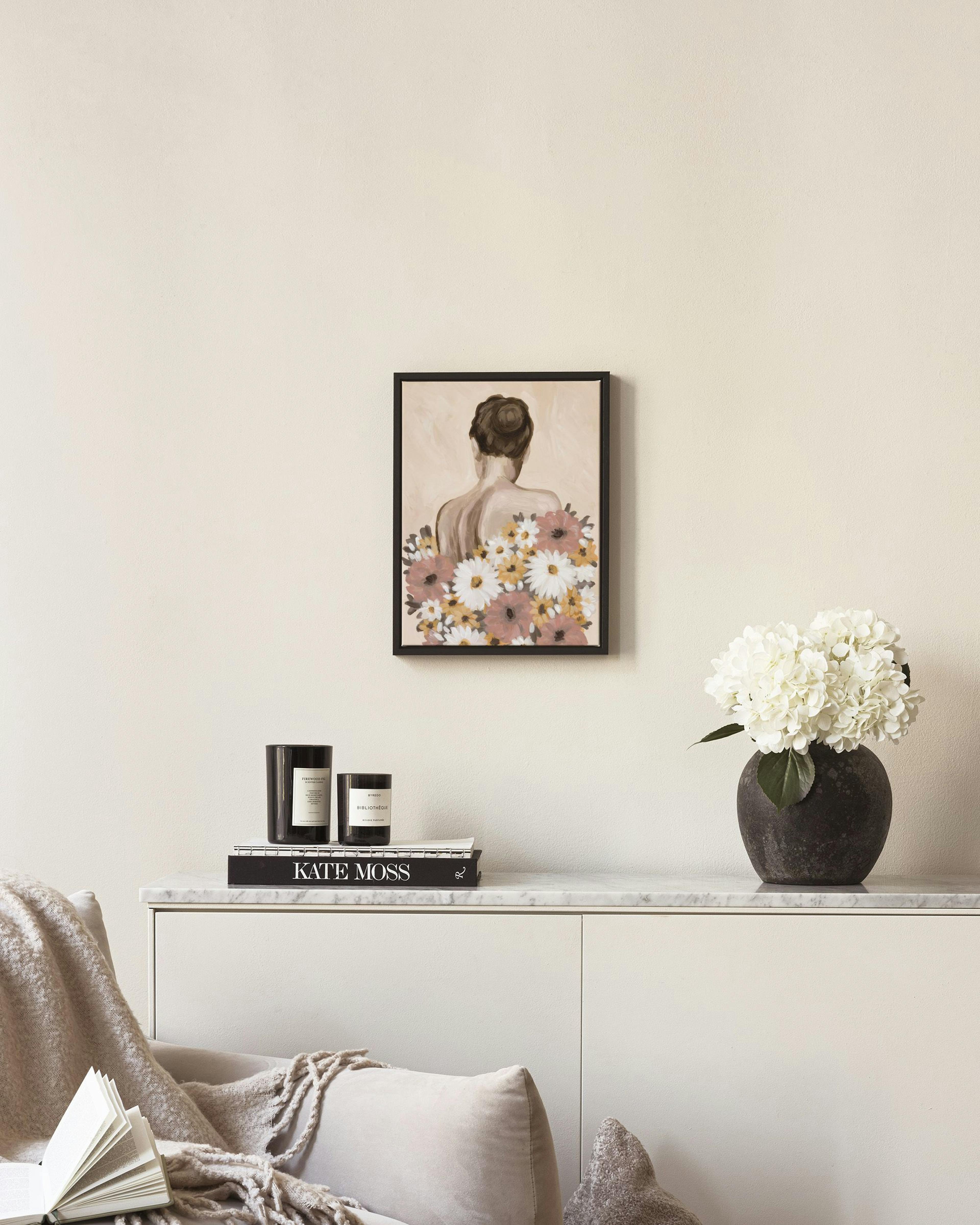 A canvas of a woman with a floral dress, hanging above a timber chair with books and a candle, next to a cane cabinet.