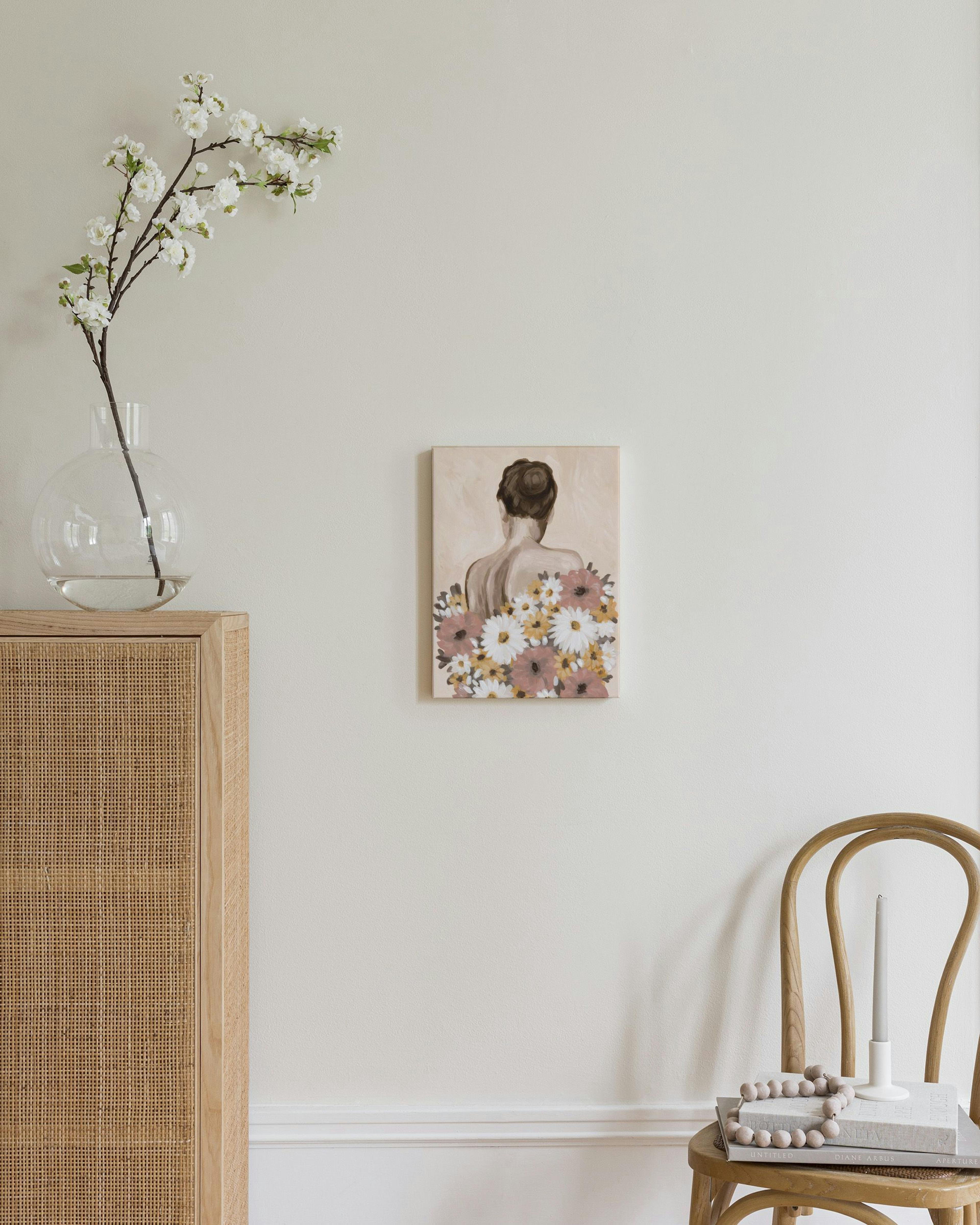 A canvas showing a womans back with a floral arrangement of white, yellow, and pink flowers, displayed on a wall in a bright roo