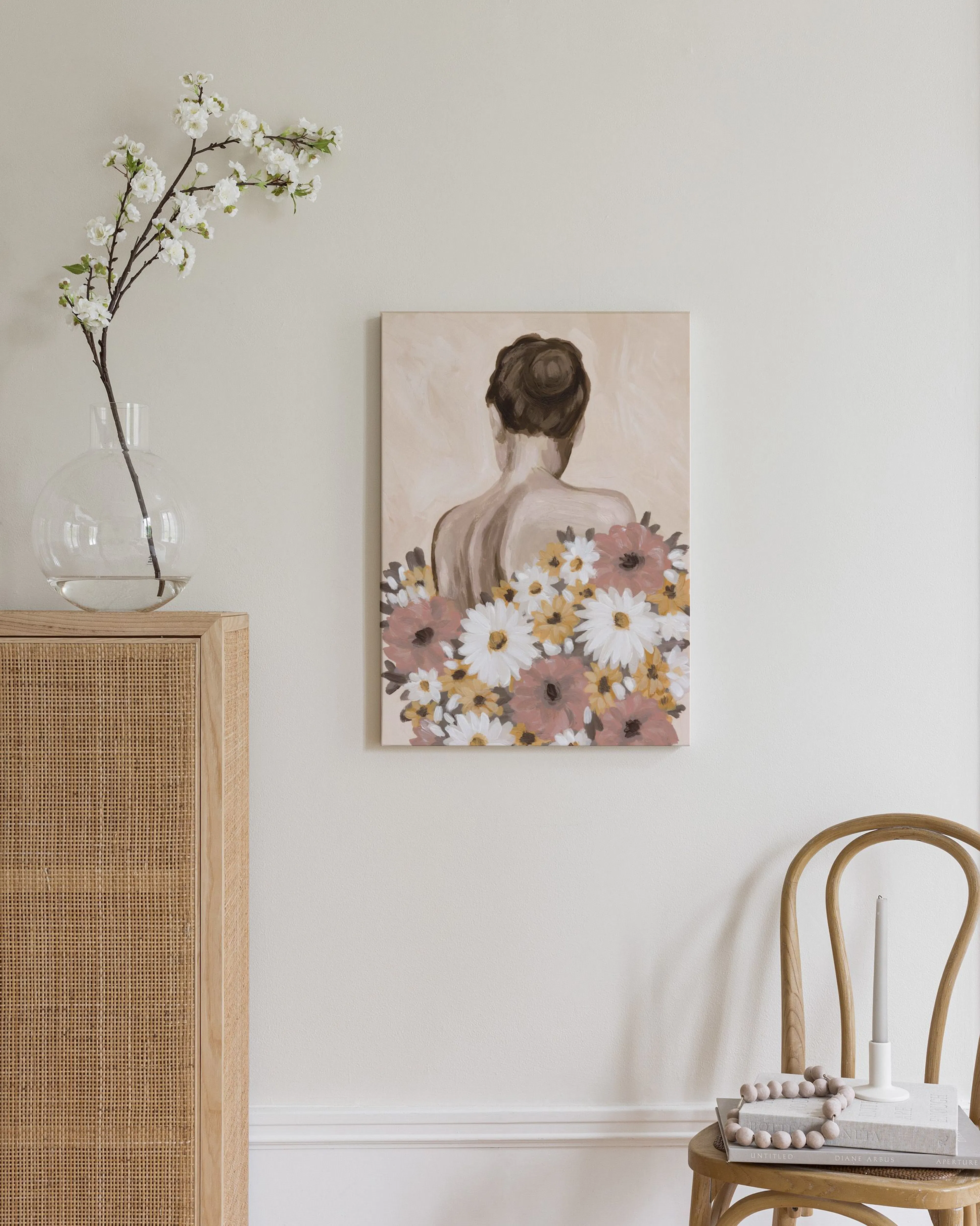 Leinwandbild einer Frau von hinten mit Blumen in Rosa, Gelb und Weiß, hängt über einem Stuhl in einem schlichten Raum.