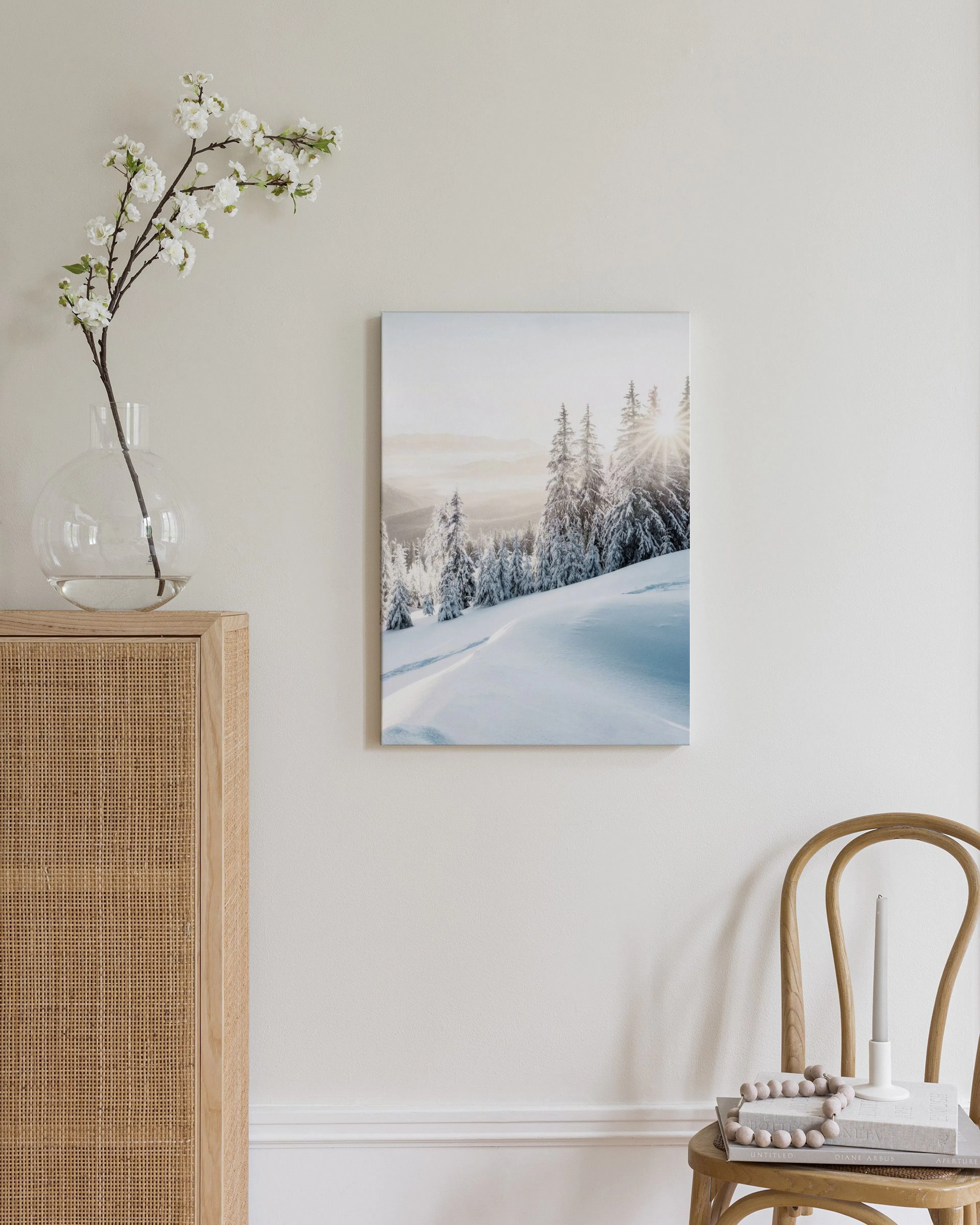 A canvas of a snowy mountain landscape with sun rays, displayed above a light wood cabinet with a lamp and books.