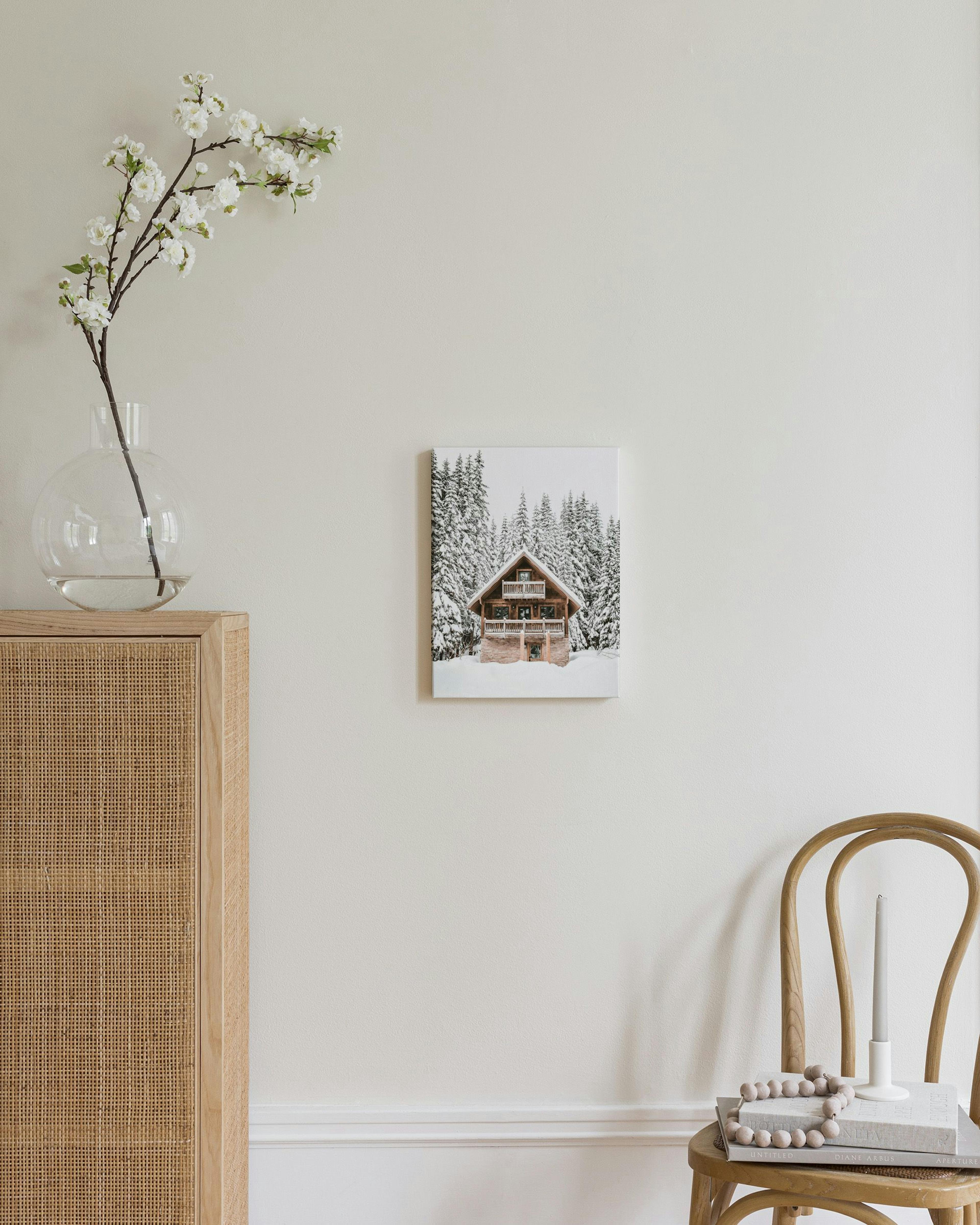 Lærred med en træhytte i snedækket skov, hængende over et flettet skab med en lampe og bøger.