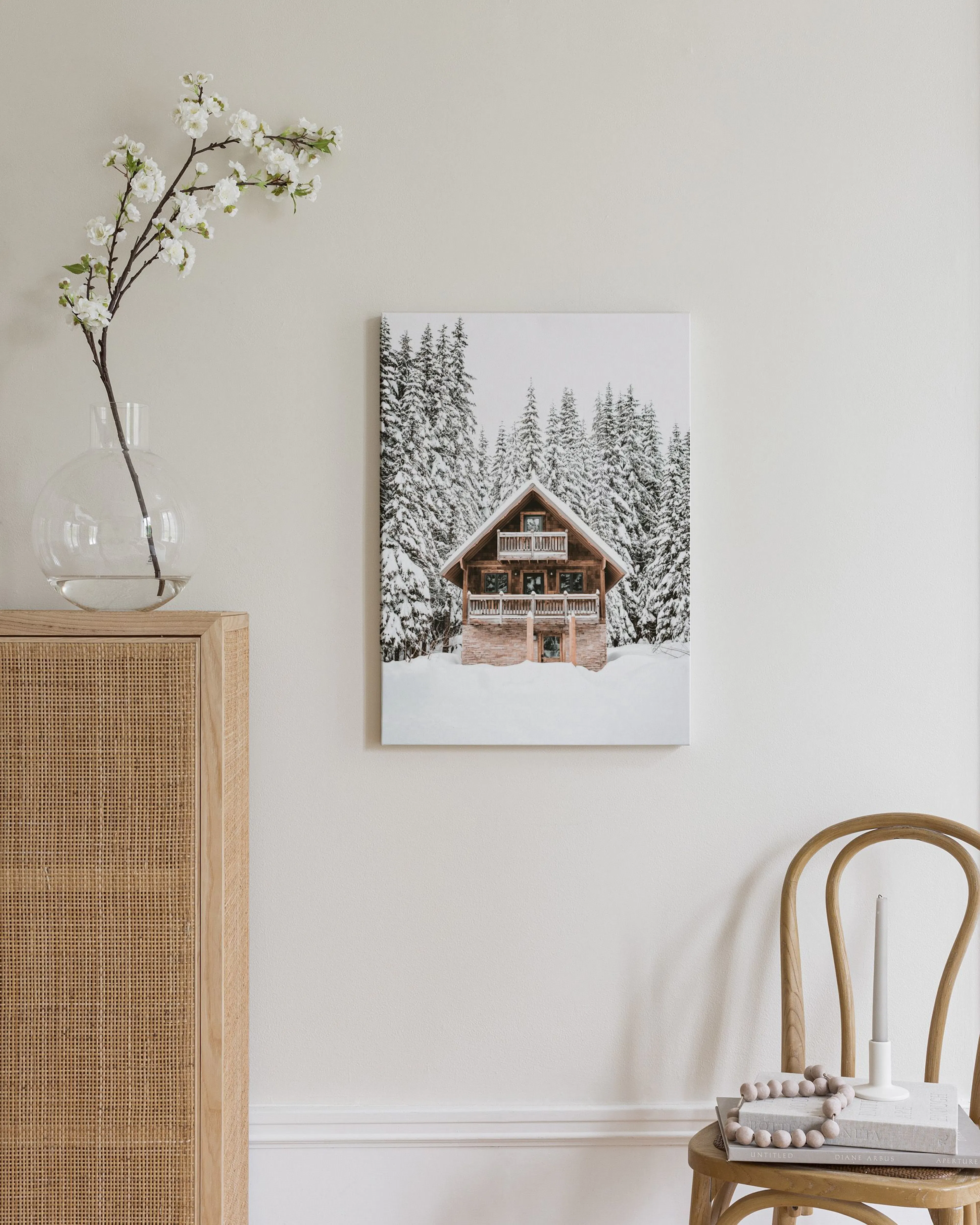 Toile dun chalet en bois enneigé au milieu de sapins, exposé sur un buffet en rotin aux côtés dune lampe et de livres.