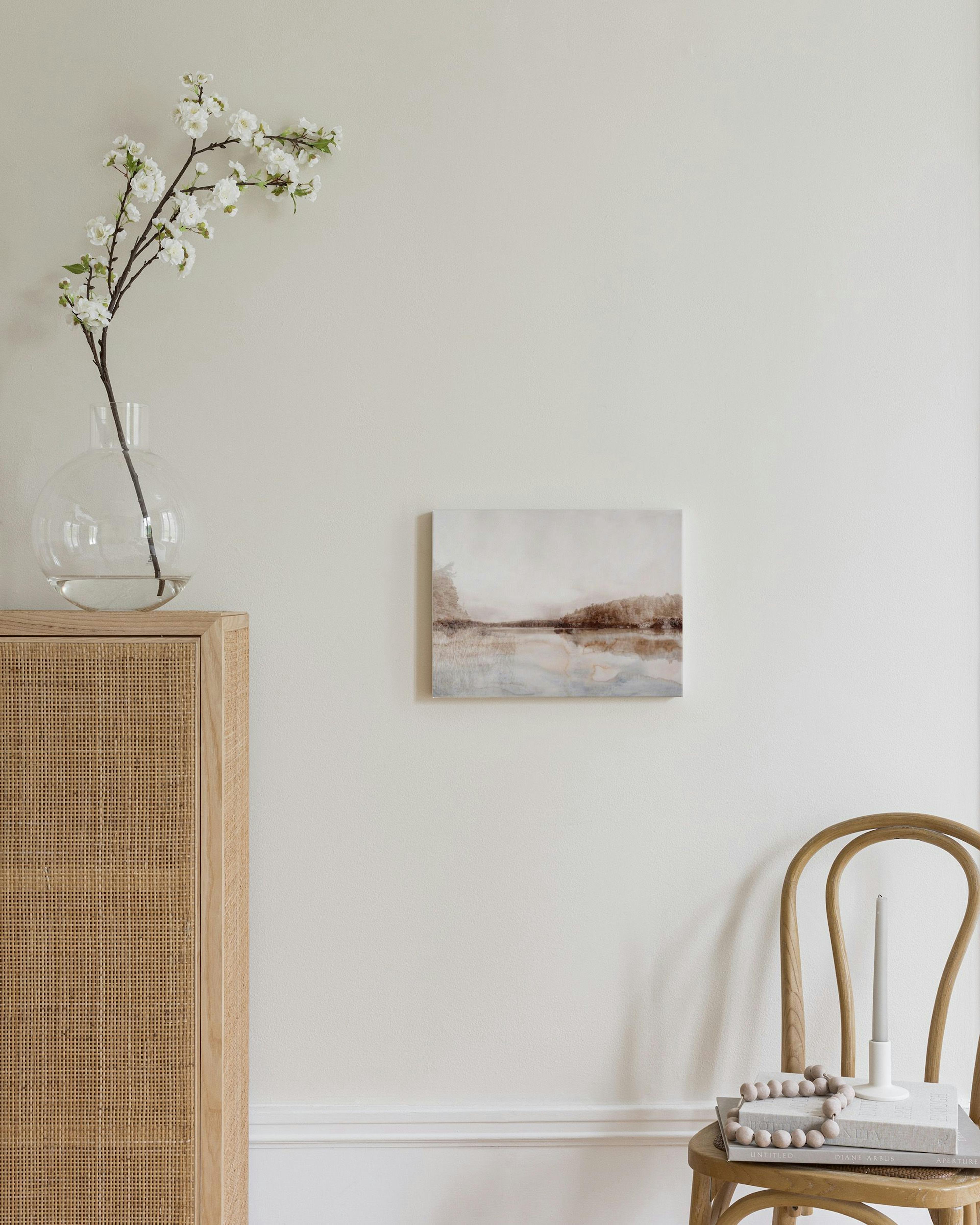 Toile abstraite de paysage lacustre avec des teintes de brun, beige et bleu pâle, suspendue au-dessus dune chaise en bois.