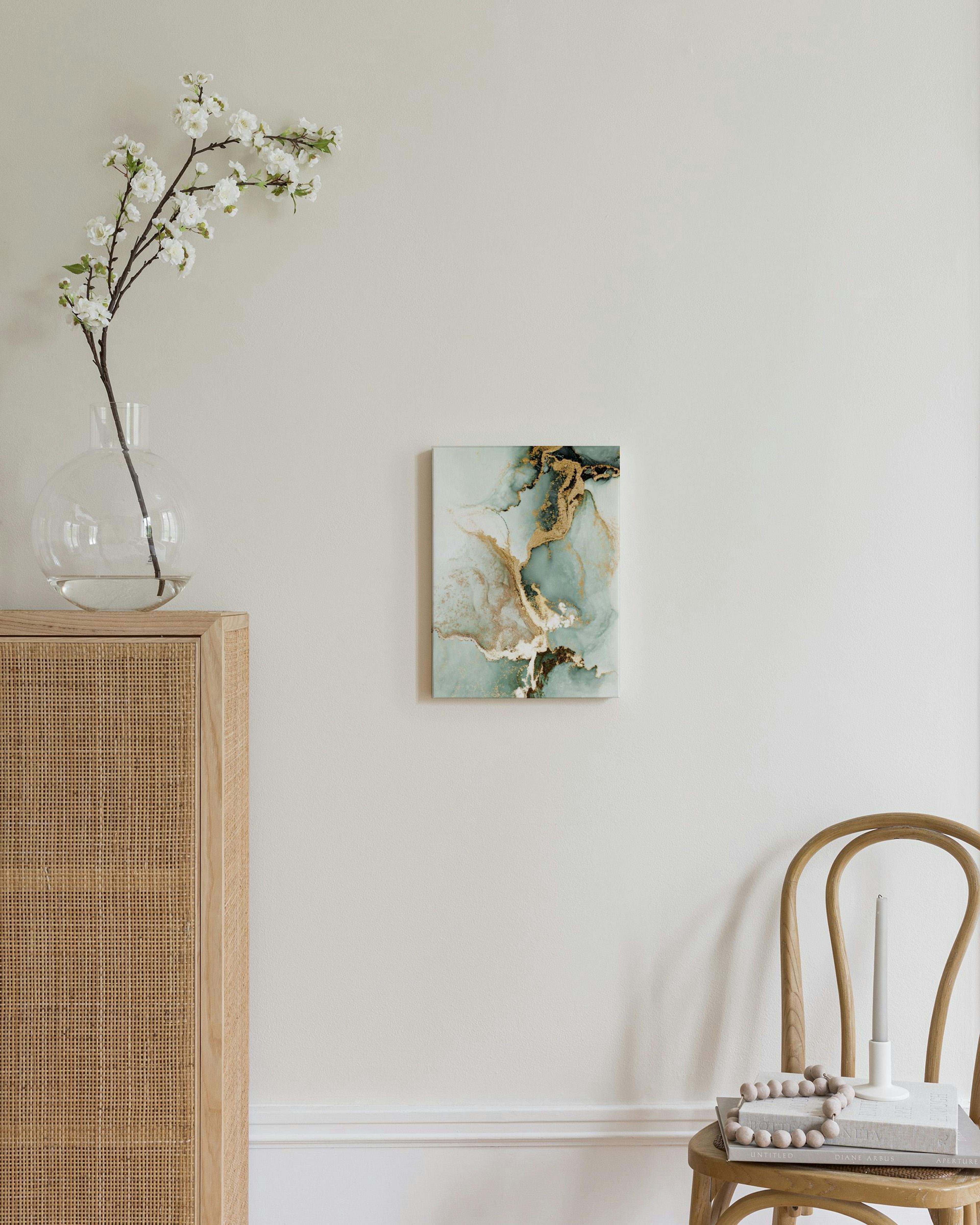 Lerretsbilde med abstrakte grønne og blå former med glitrende gull og hvite detaljer, henger over et trebord.