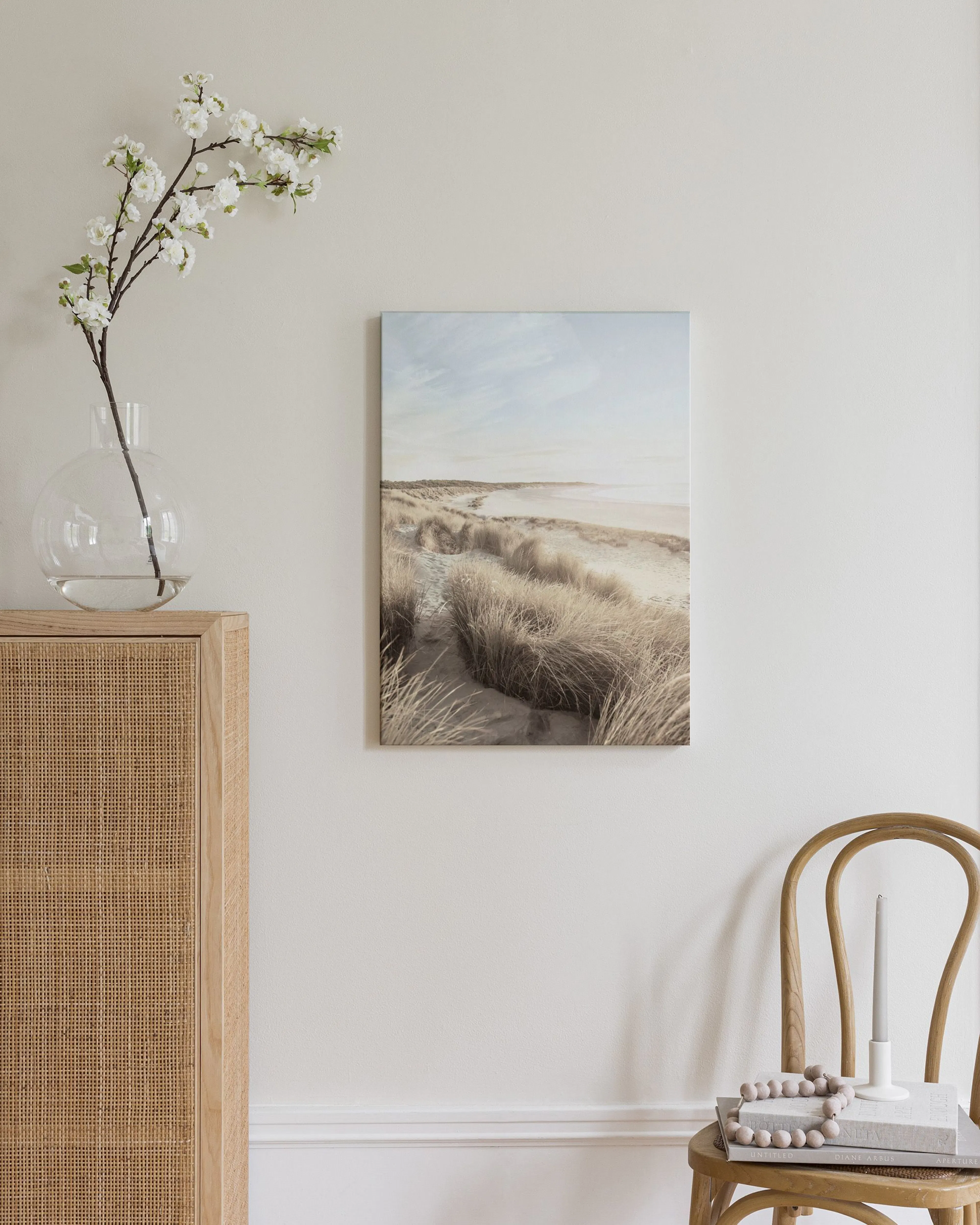 Toile de plage avec dunes et herbes sèches, ciel clair, affichée au-dessus dun meuble en rotin dans une chambre.