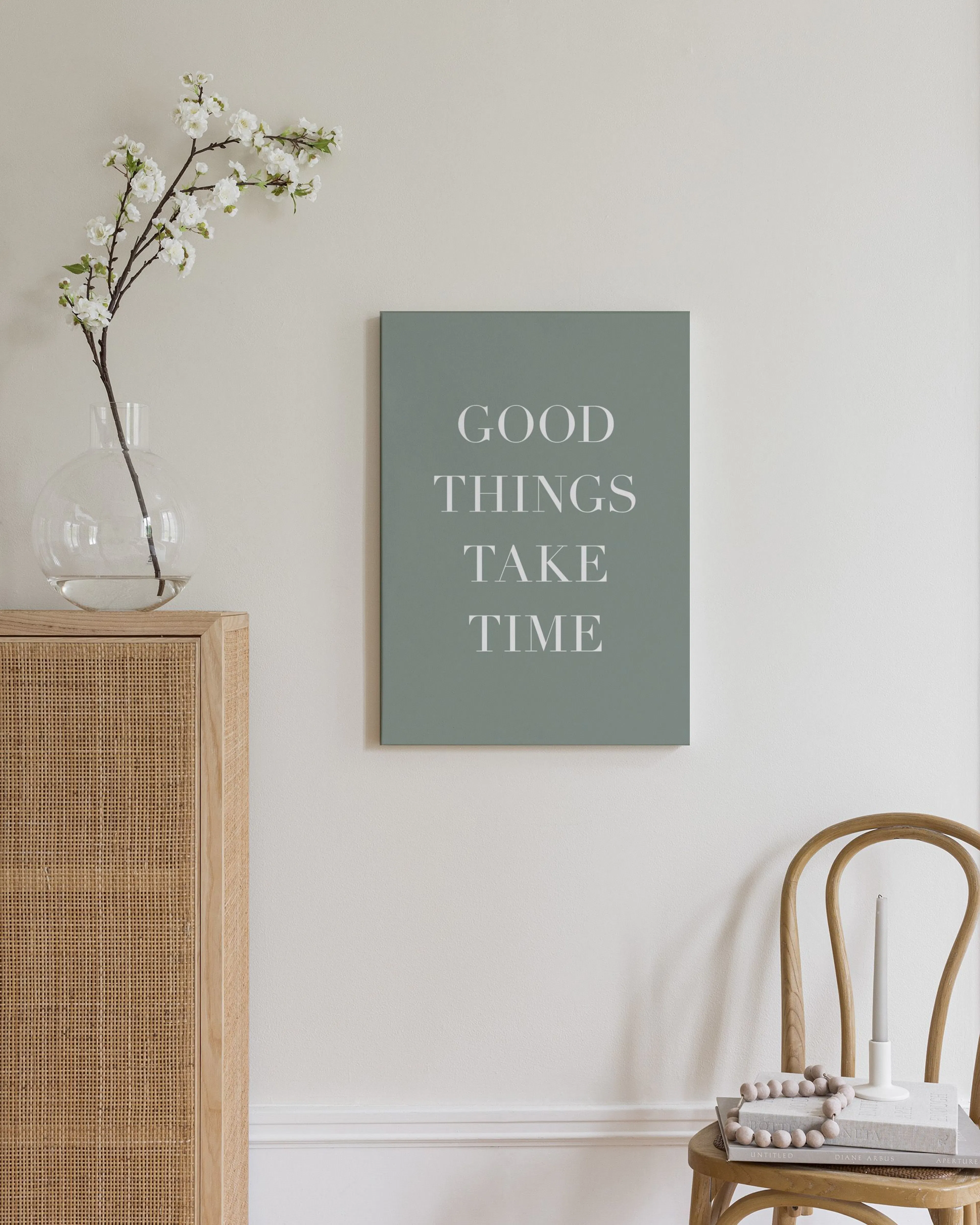 Canvas met witte tekst GOOD THINGS TAKE TIME op een sagegroene achtergrond, hangend boven een houten dressoir in een slaapkamer.