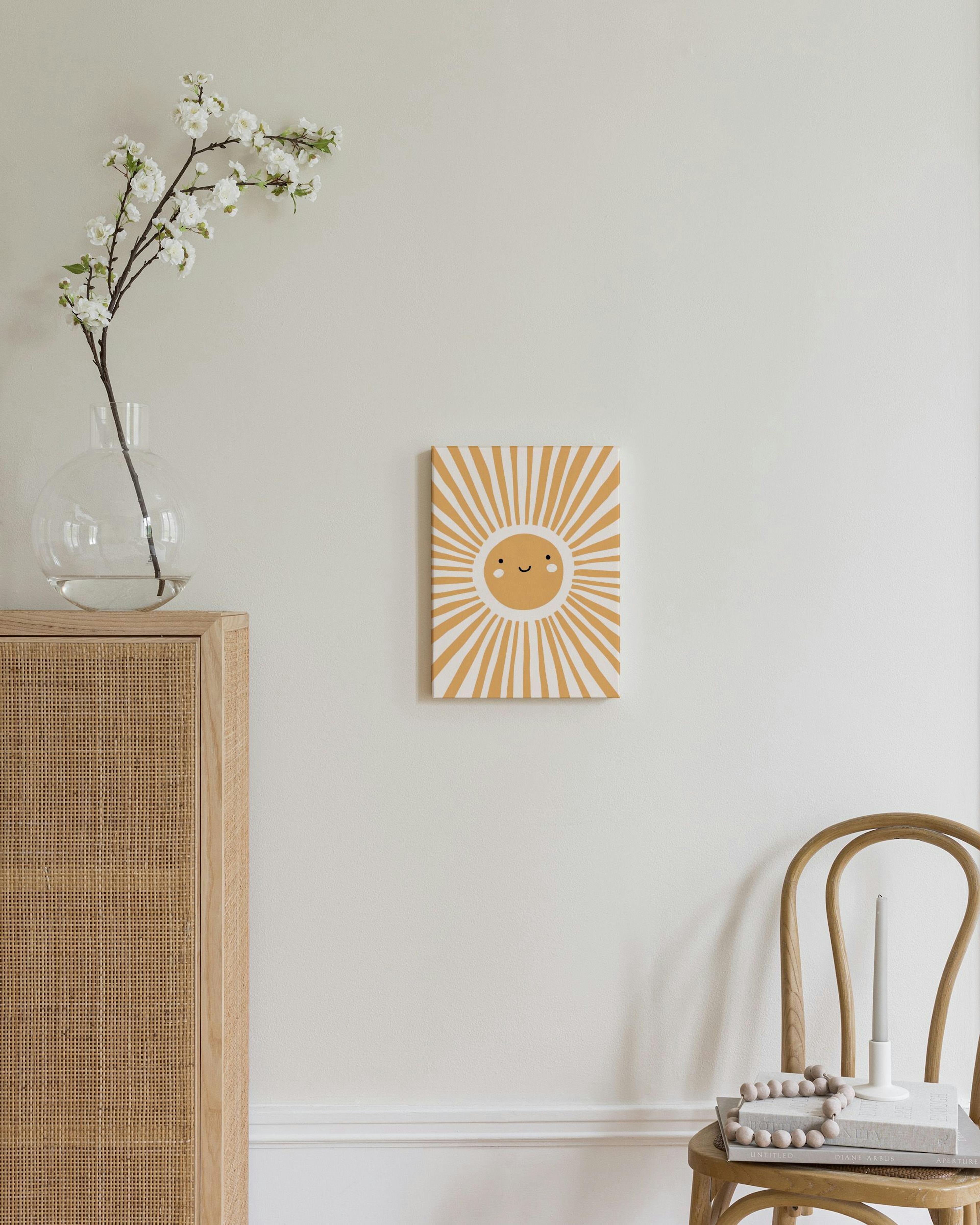 Toile murale soleil souriant jaune et blanc, au-dessus dune commode en bois et rotin, avec lampe et livres.