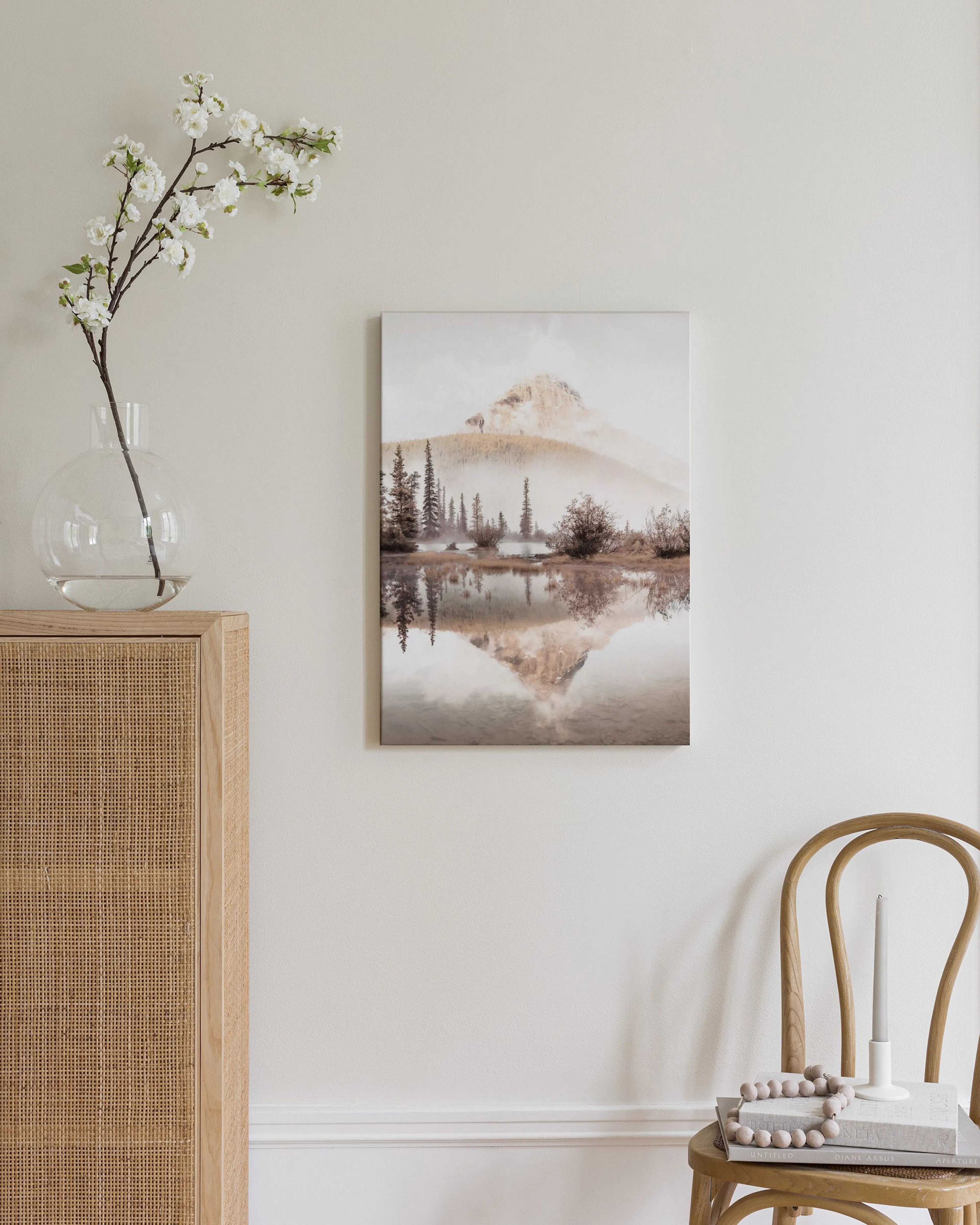A misty mountain landscape with pine trees and a reflective lake, displayed above a wooden cabinet with a lamp and books.