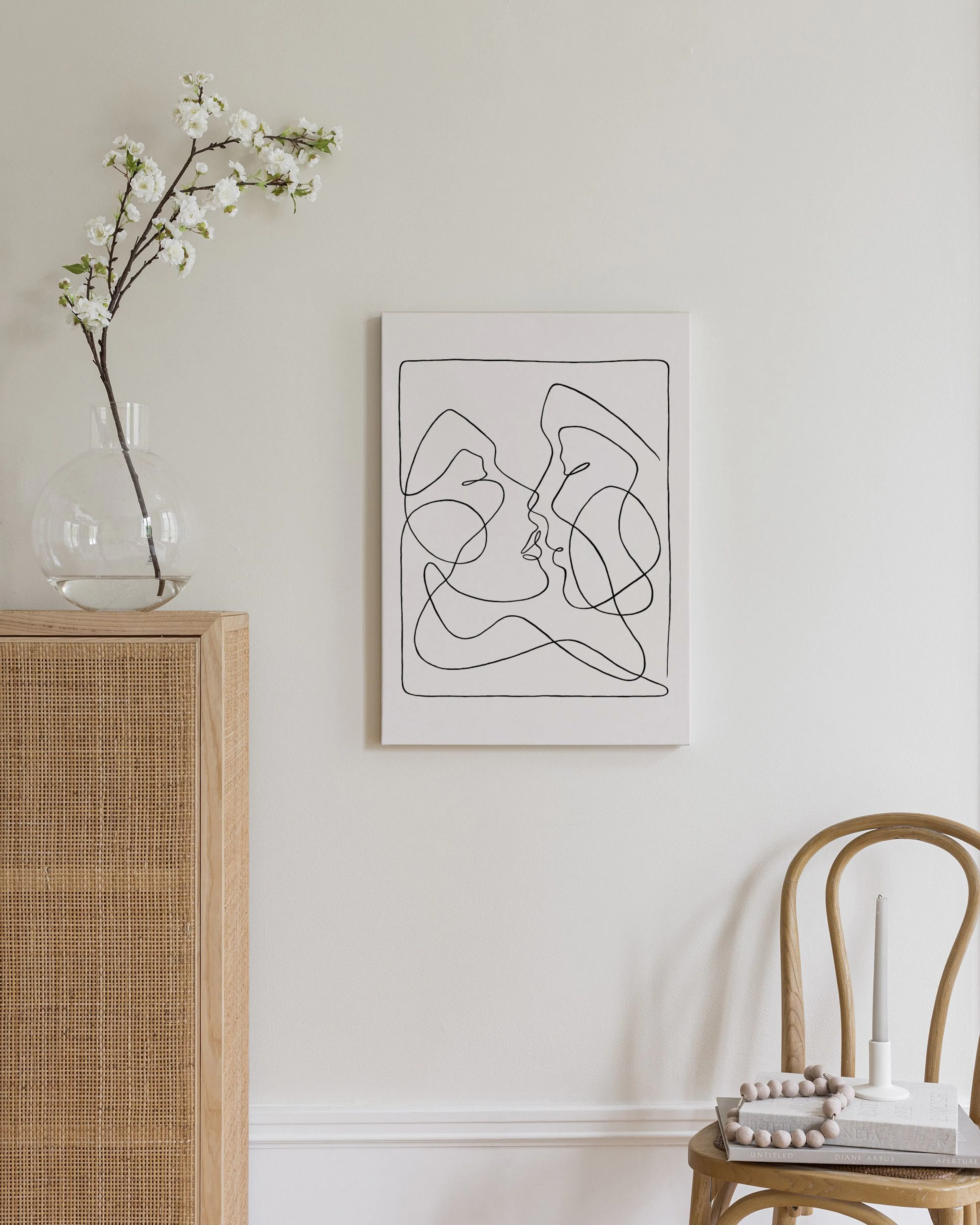 Toile murale minimaliste dune ligne noire continue formant deux visages sembrassant, au-dessus dune commode en rotin et bois cla
