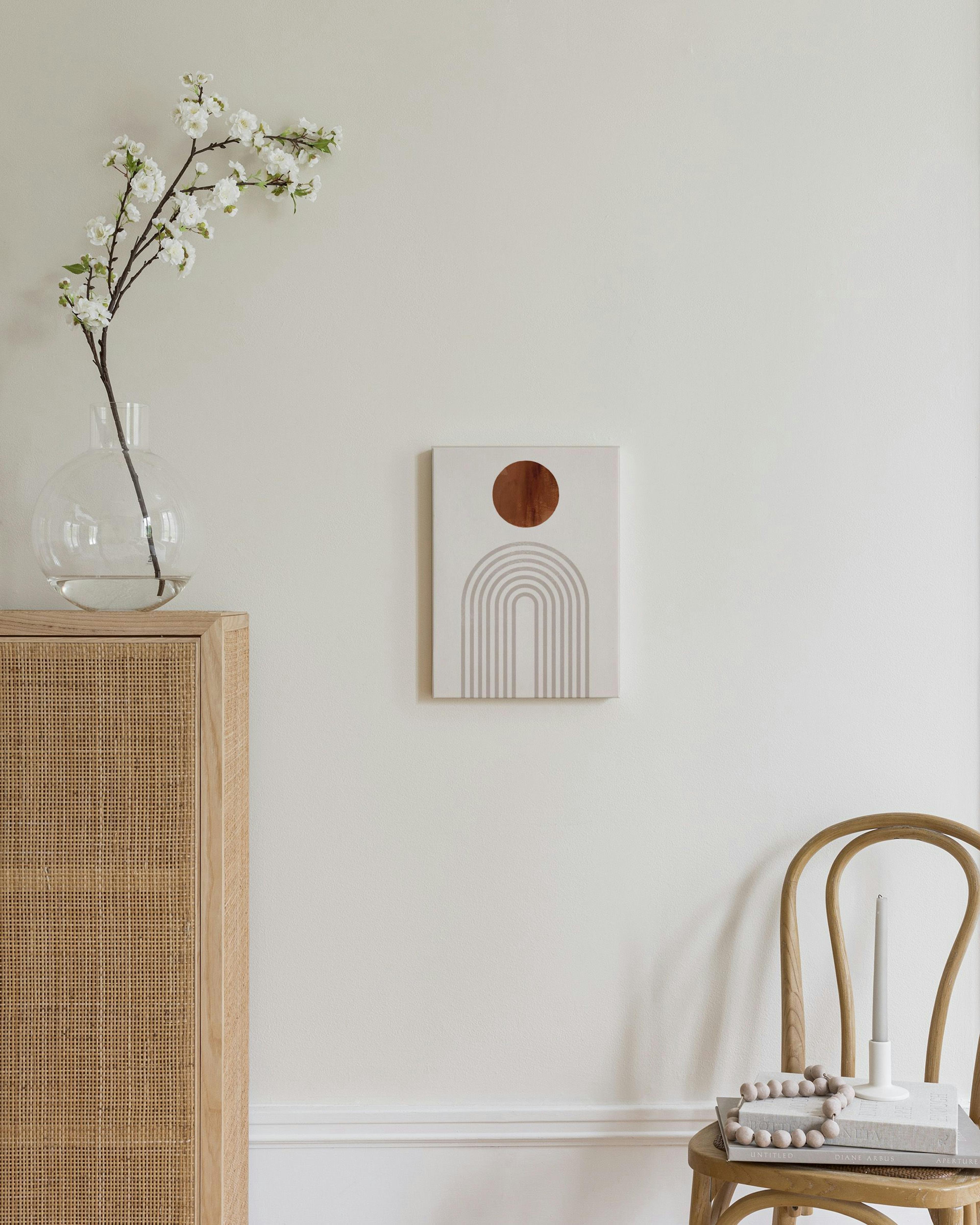 A canvas with a rust-coloured circle and grey arched lines, hanging above a natural wood and wicker cabinet in a bedroom.