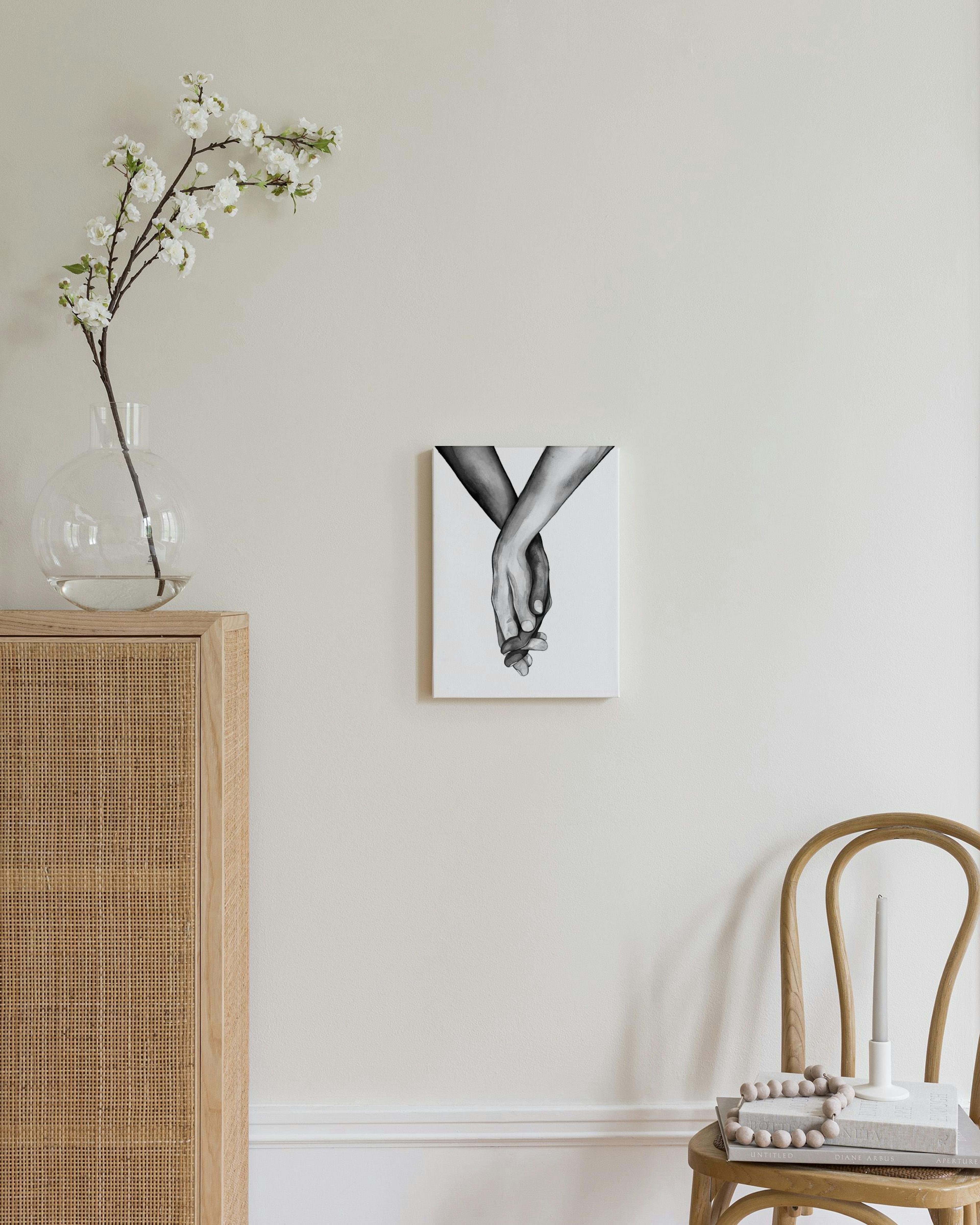 Kanvaasi, jossa mustavalkoinen piirros toisiinsa kietoutuneista käsistä, ripustettuna beigelle seinälle makuuhuoneessa.