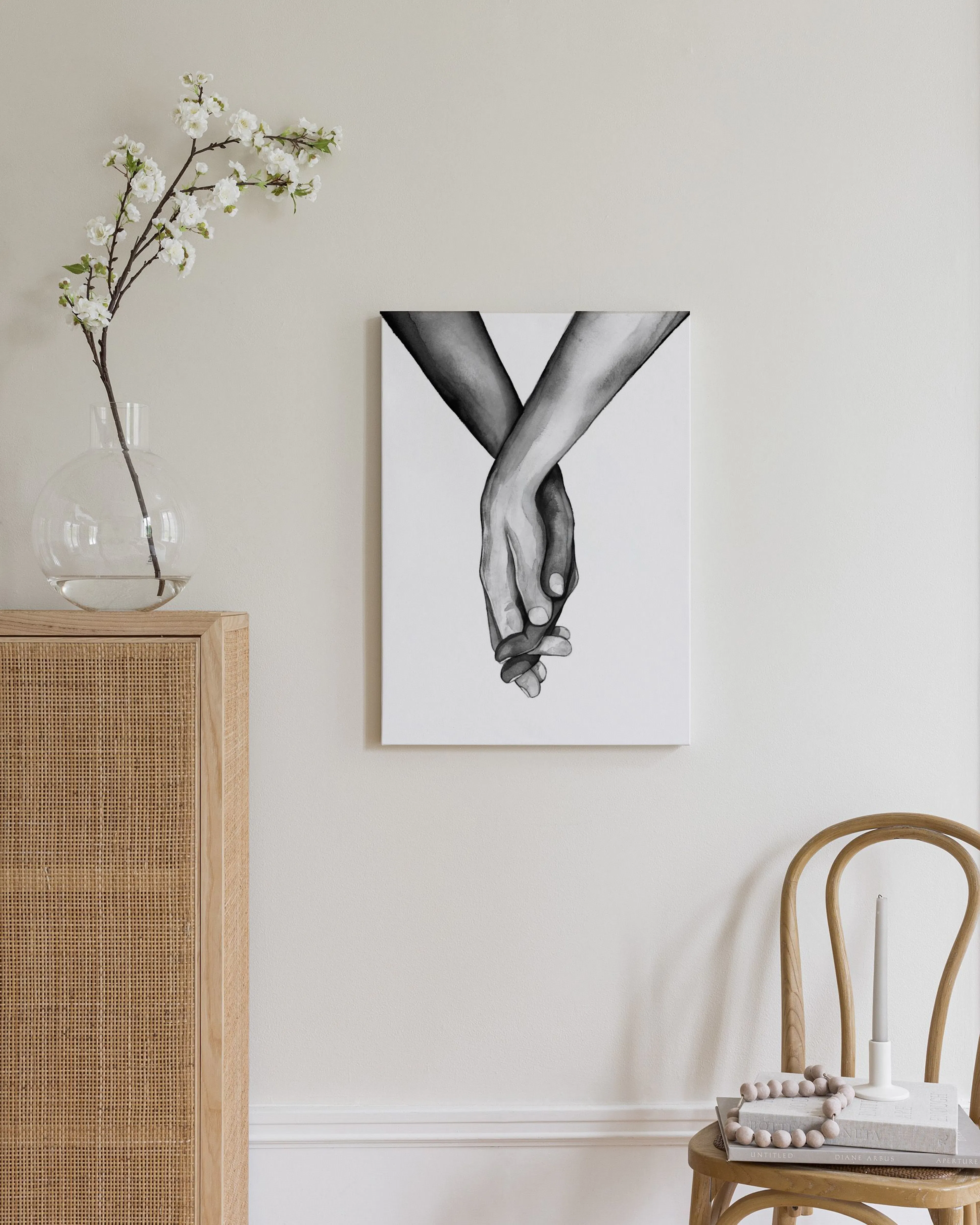 A black and white canvas of two hands holding, displayed above a wooden dresser with a lamp and decor.