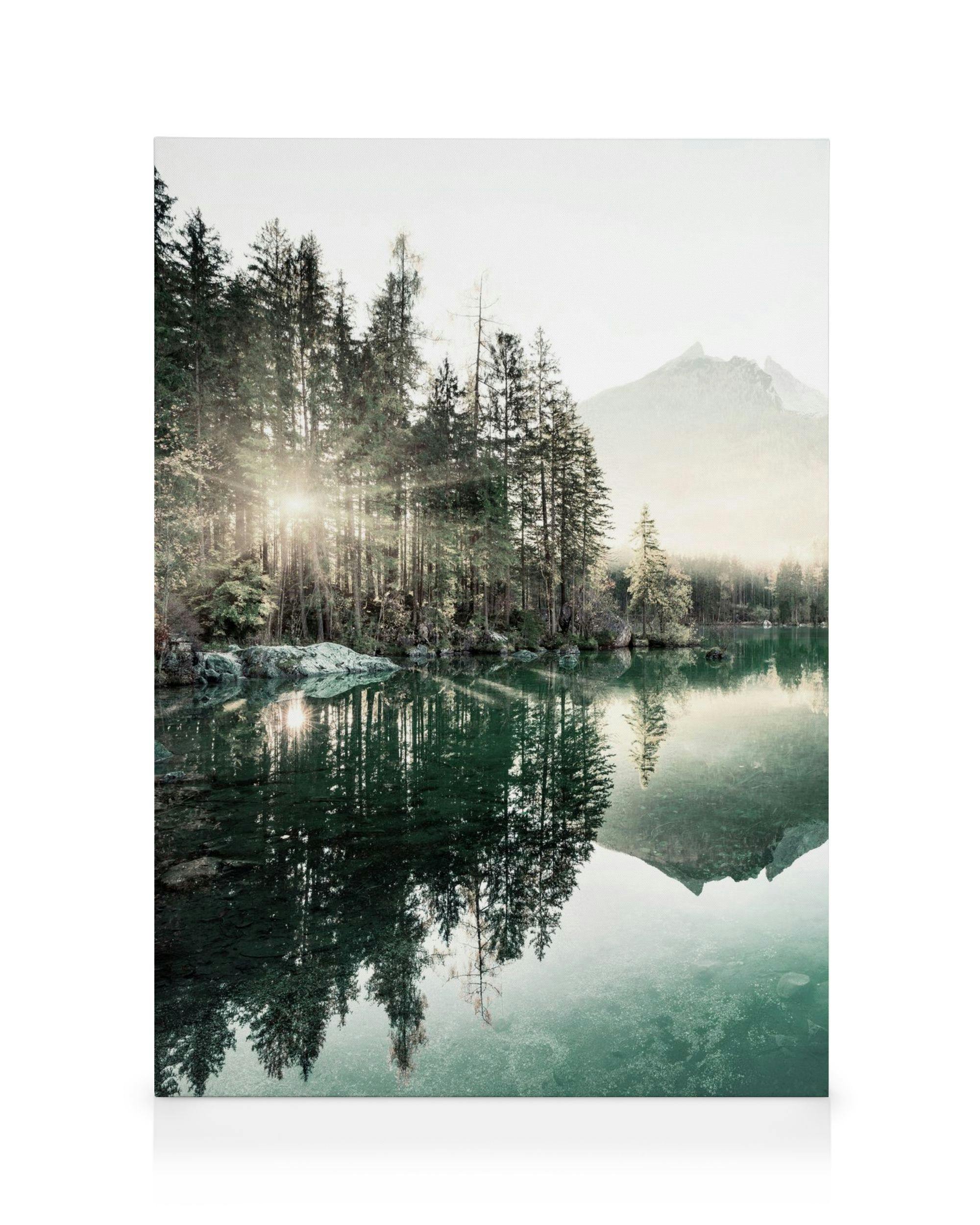 Leinwandbild mit Sonnenstrahlen durch einen Nadelwald, der sich in einem grünen See mit Berglandschaft spiegelt.