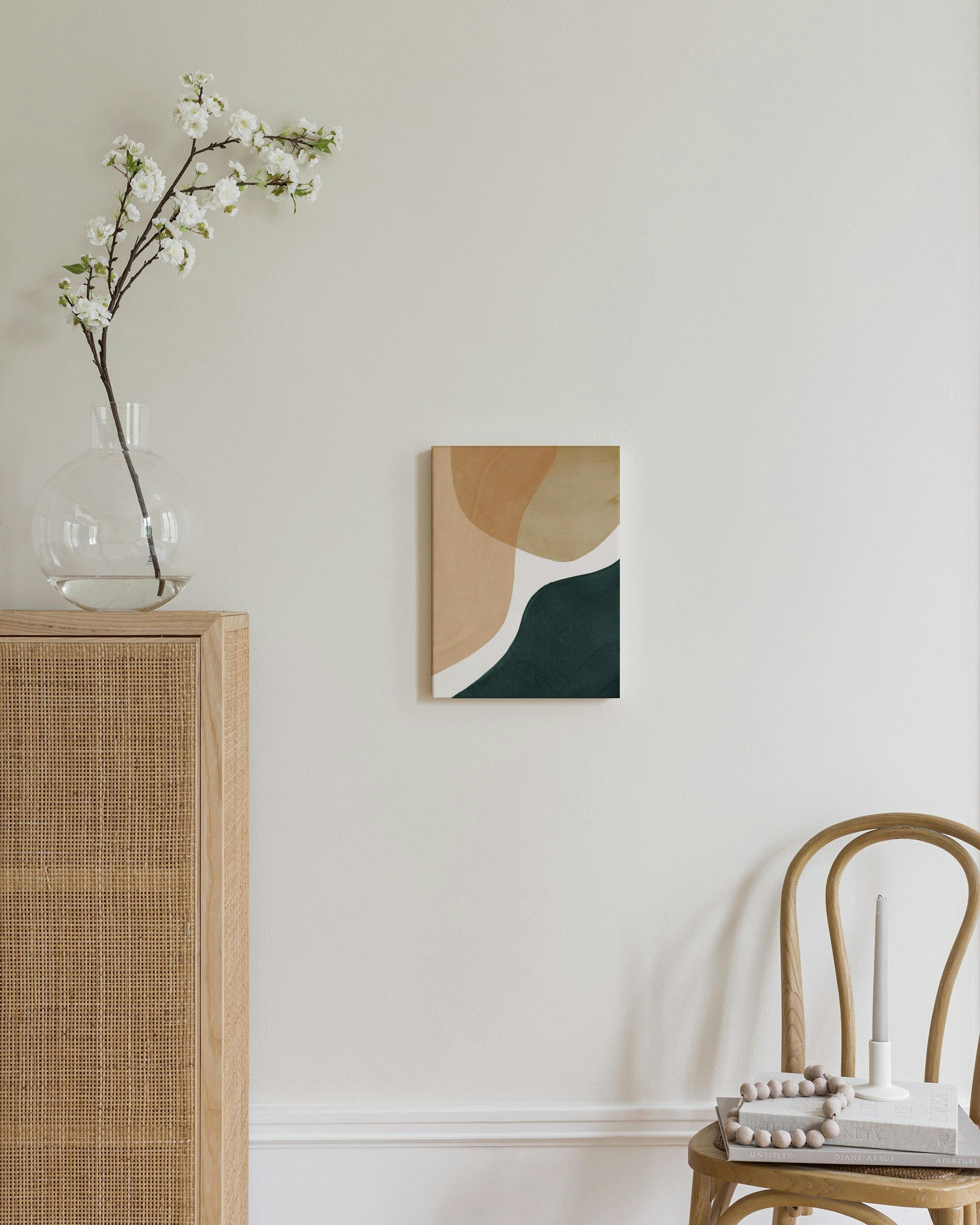 A canvas with abstract organic shapes in beige, brown, white, and dark green, displayed above a rattan cabinet.