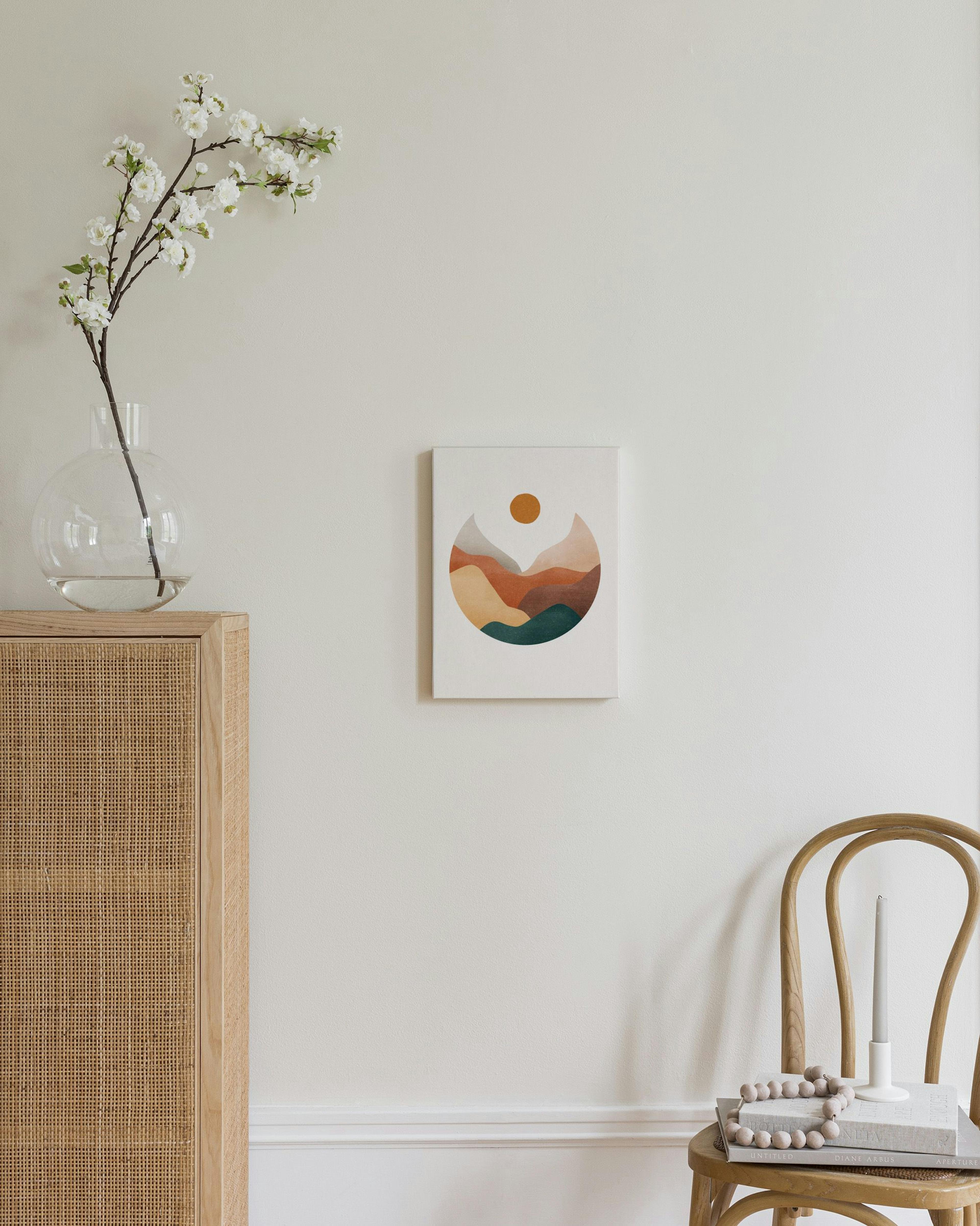 Et lerretsbilde med et abstrakt landskap i jordfarger og en sol, henger over et møbel i et lyst rom.