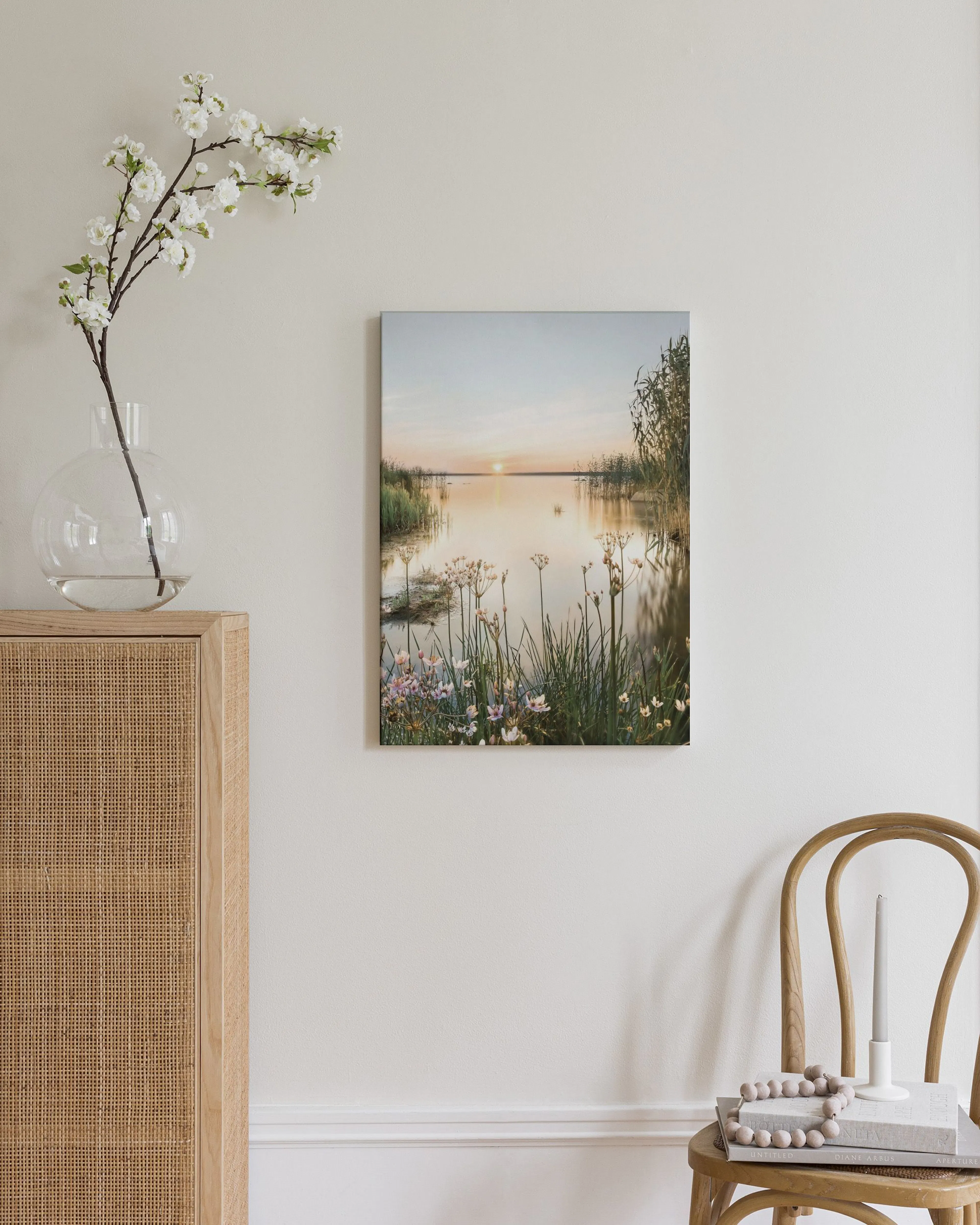 Lienzo de un atardecer dorado sobre un lago con vegetación exuberante, exhibido sobre una cómoda de madera con cojines blancos.