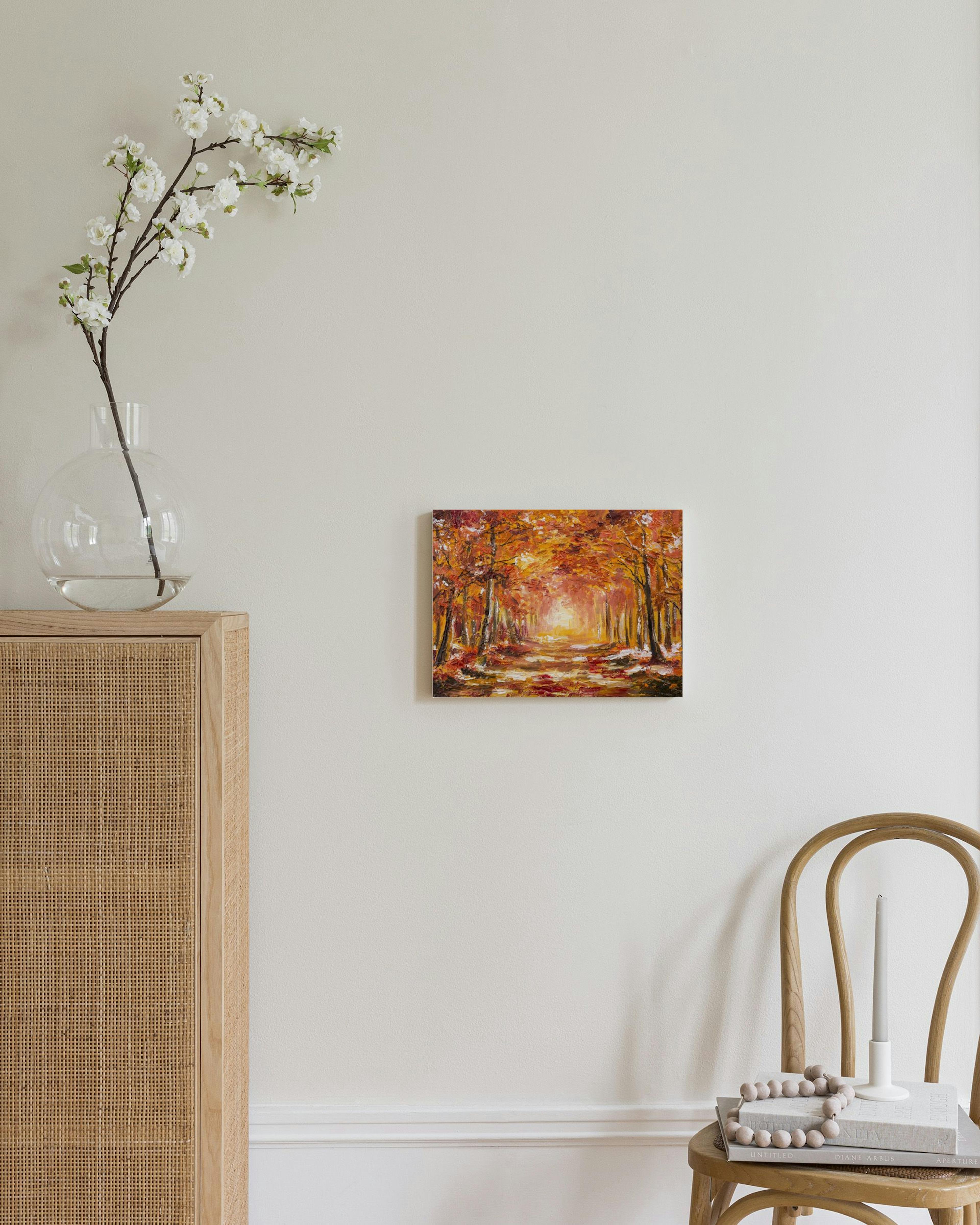 Lienzo de un sendero otoñal en un bosque con árboles de hojas rojizas y anaranjadas, colgado sobre un mueble de madera.