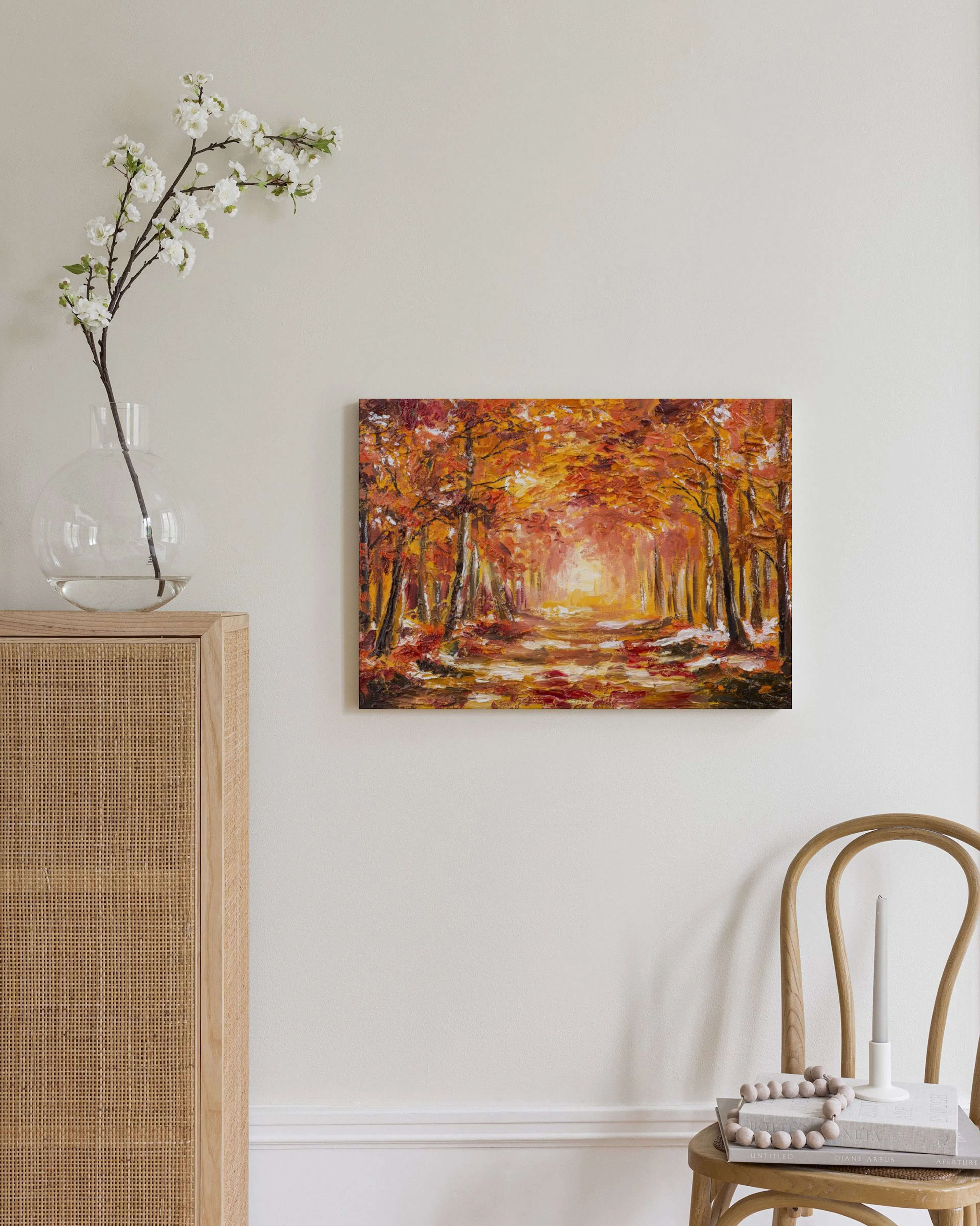 Toile dun chemin forestier en automne aux couleurs chaudes, accrochée dans un salon moderne à côté dun meuble en rotin clair.