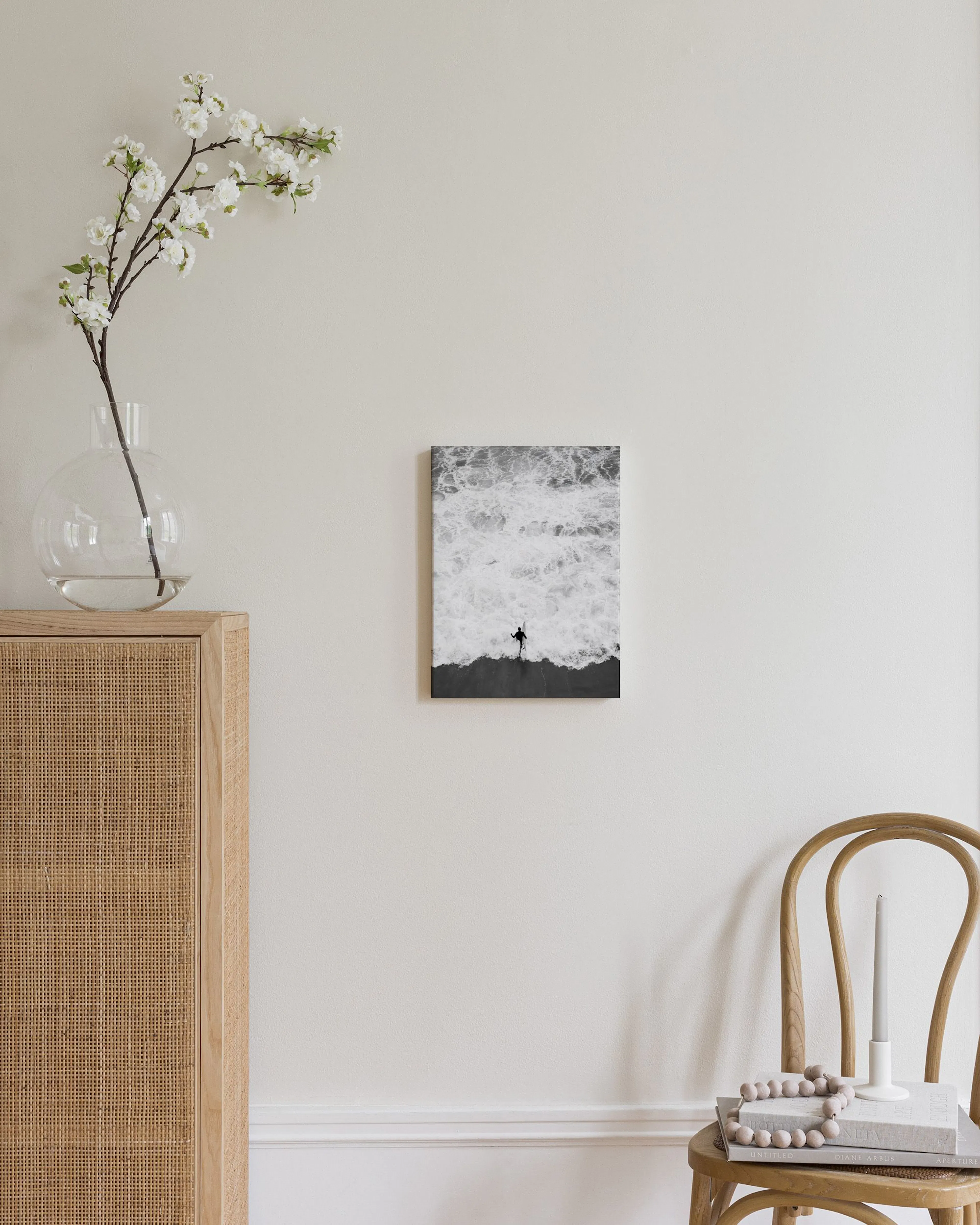 Black and white print of a surfer with a board on a dark sand beach with foamy ocean waves.