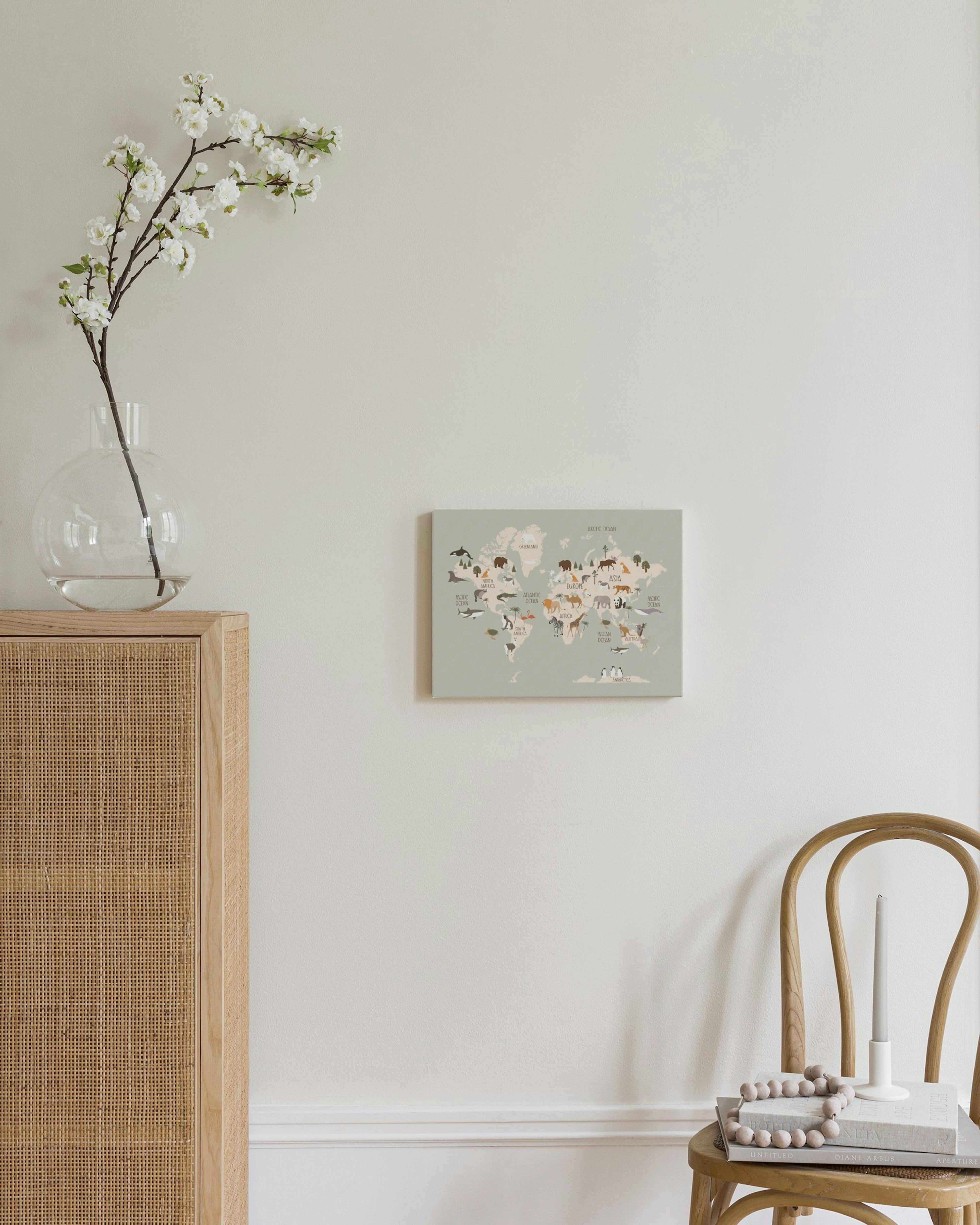 A canvas of a world map in muted green with illustrated animals, hanging in a bright room above a white ottoman.