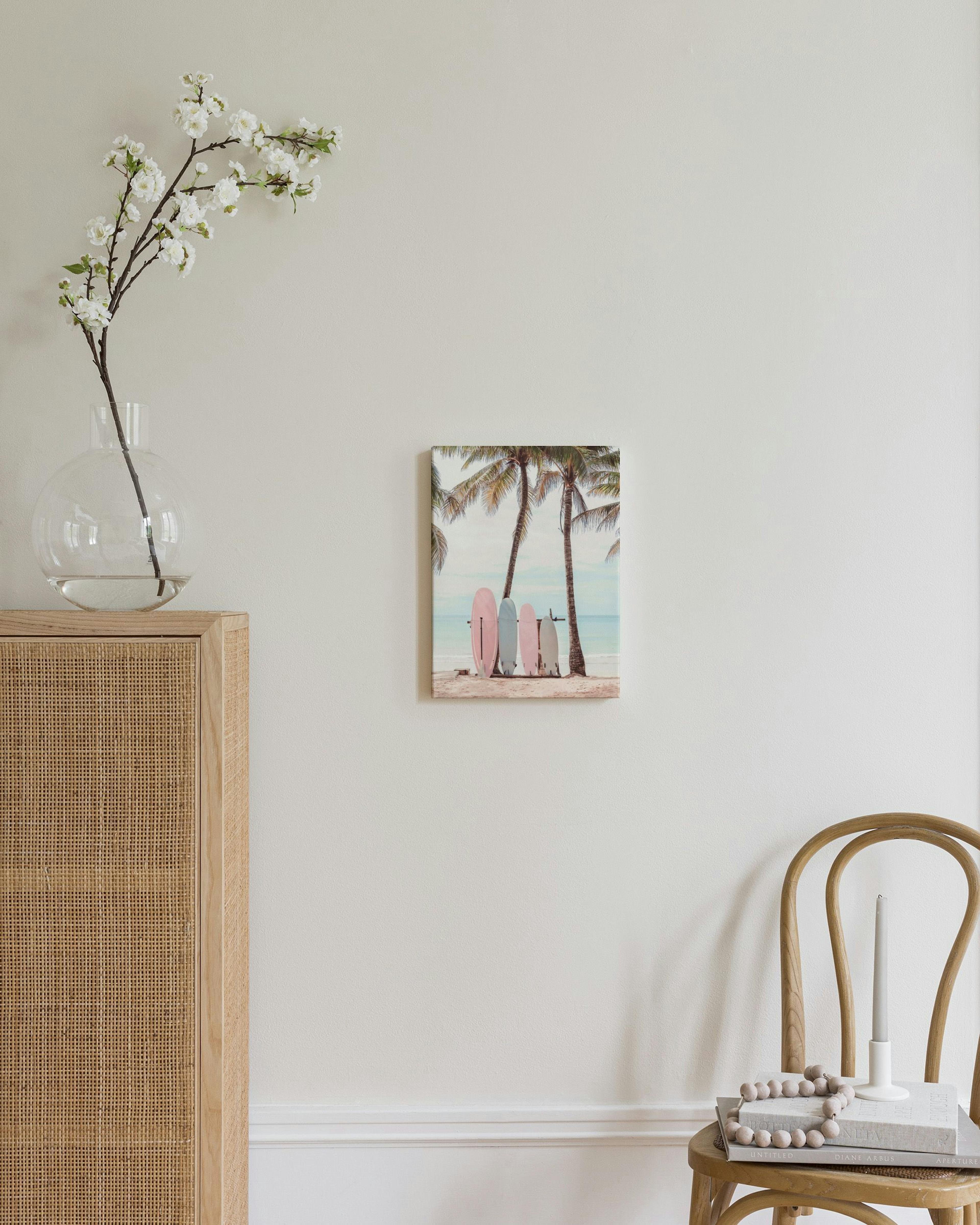 Leinwandbild mit rosa und hellblauen Surfbrettern unter Palmen am Strand, an einer beigen Wand im Wohnzimmer.