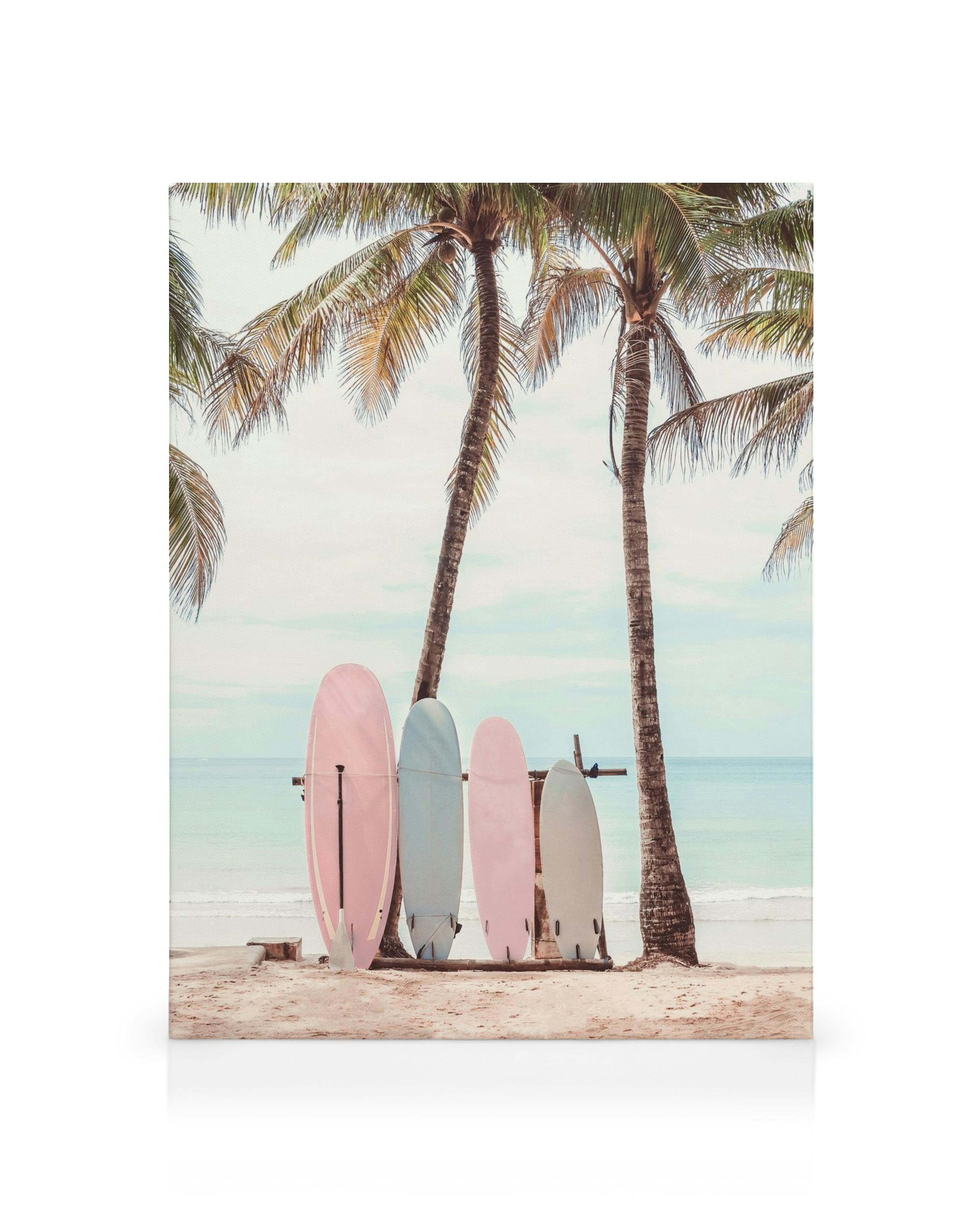Leinwandbild mit pastellfarbenen Surfbrettern unter Palmen am Strand mit Meer im Hintergrund.