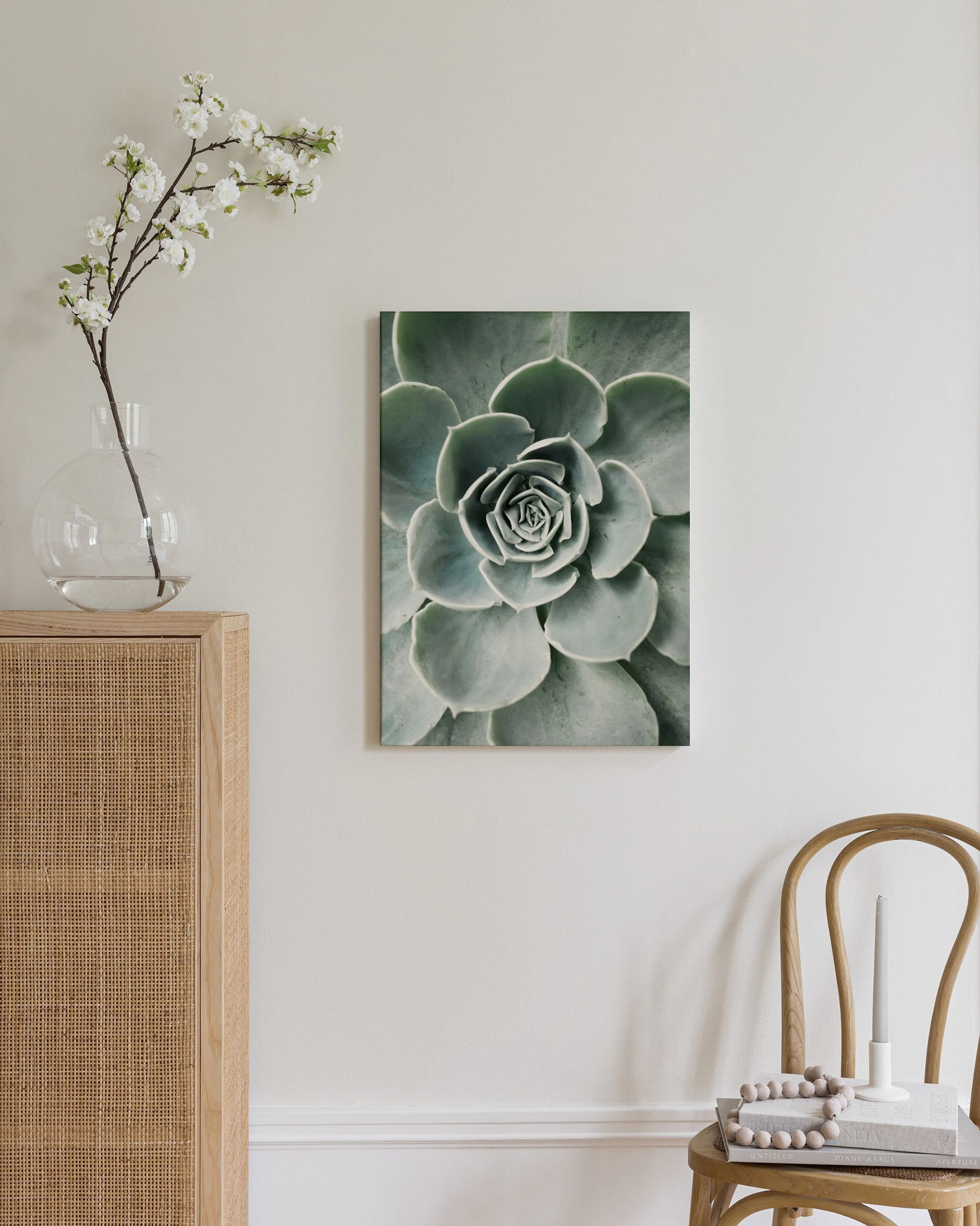 A canvas of a green succulent rosette hanging on a beige wall above a white ottoman with a book and blanket.