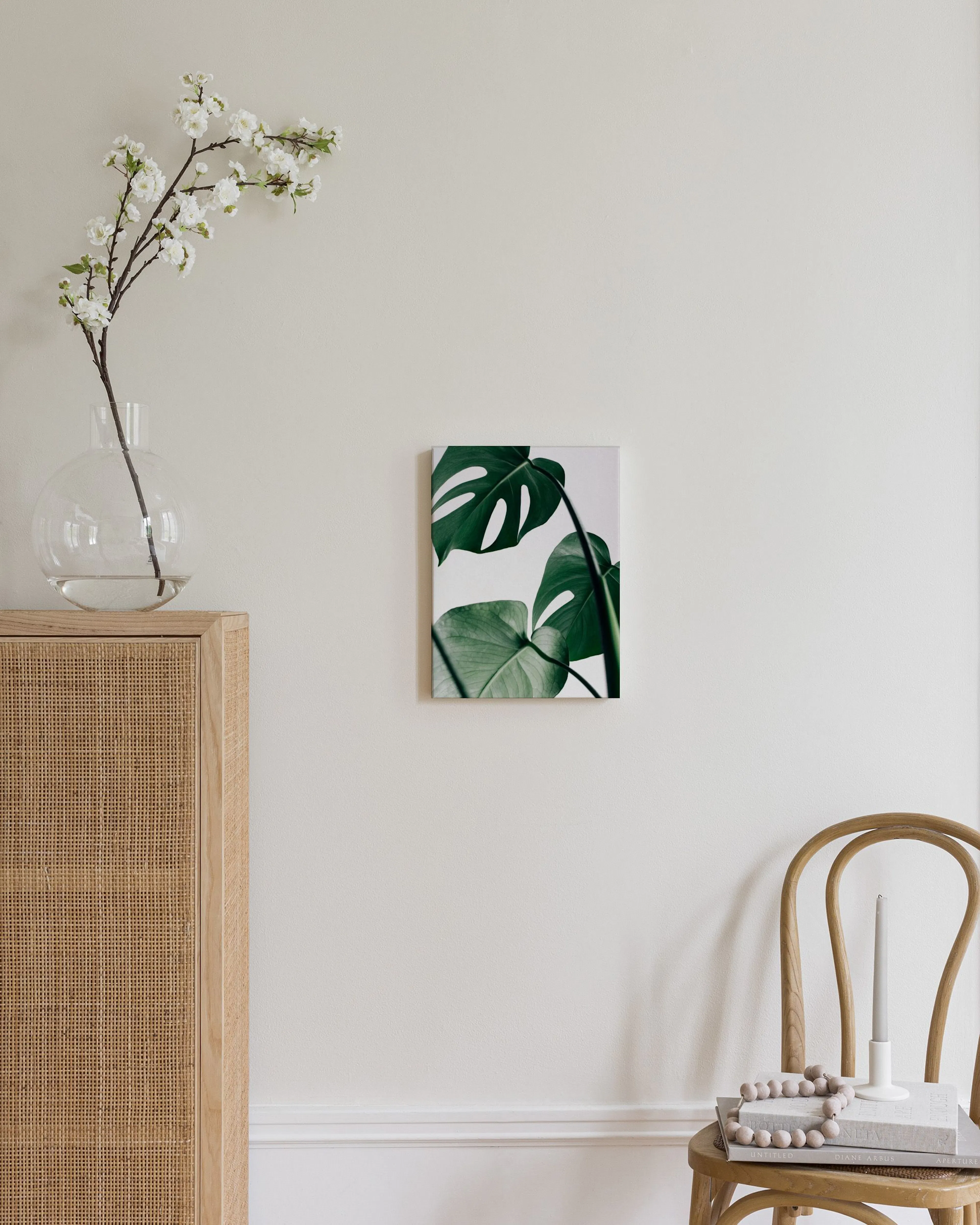 Lienzo con hojas de monstera verde sobre fondo blanco, colgado en una pared beige junto a un jarrón y un puf.