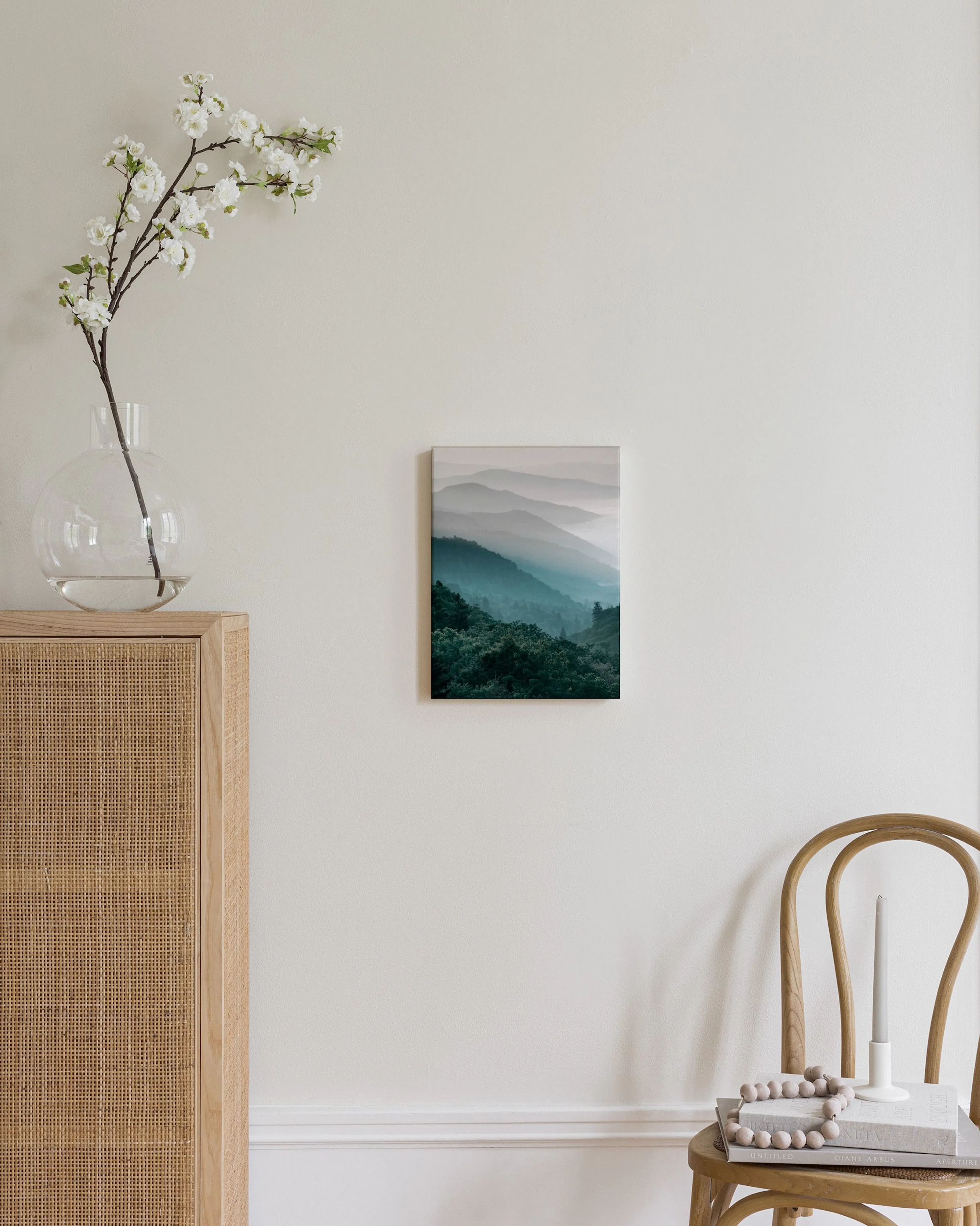 Toile murale de montagnes verdoyantes et brumeuses, accrochée dans un salon moderne au-dessus dun pouf blanc.
